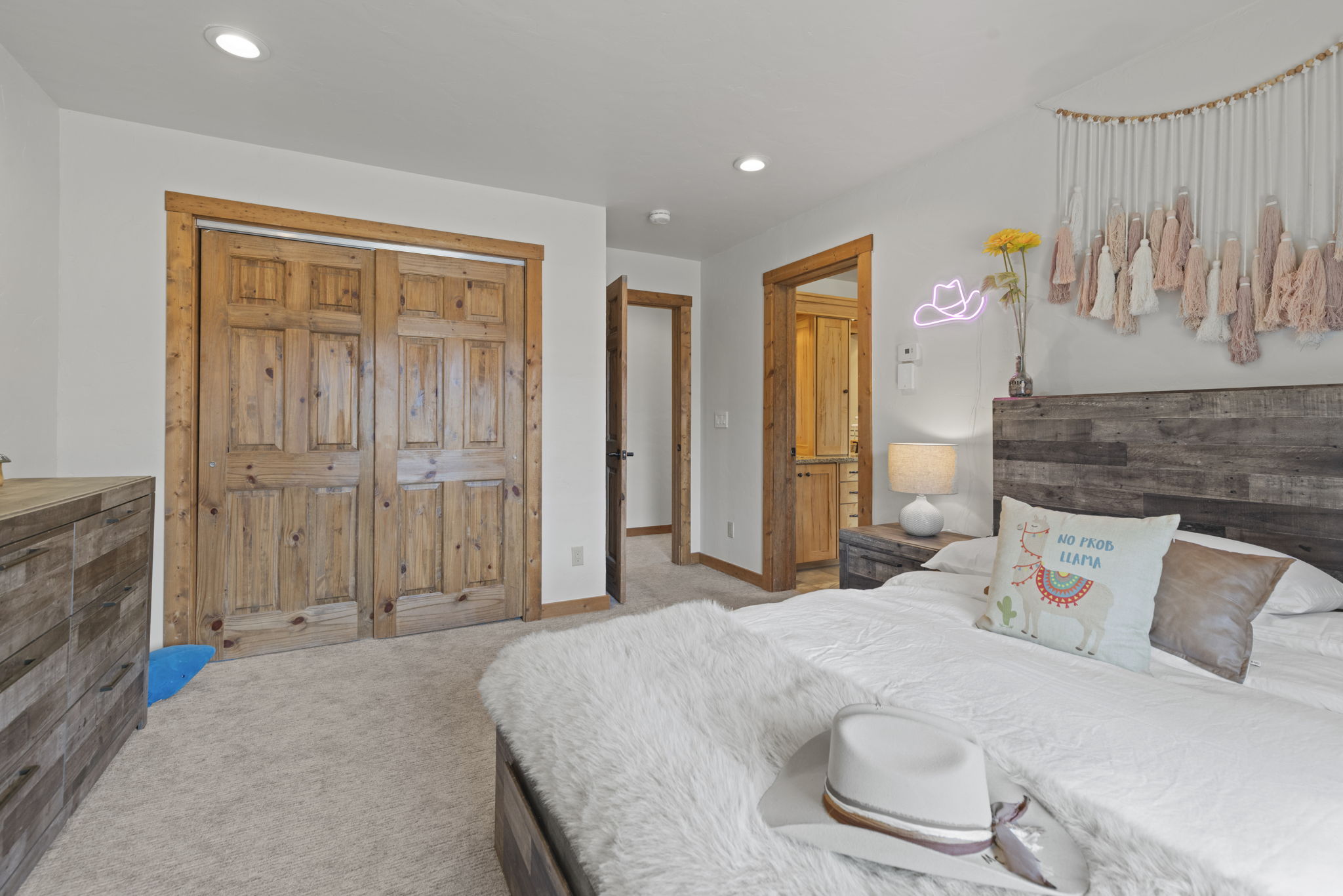 This cozy bedroom features a rustic aesthetic with a wood-paneled headboard, matching wooden closet doors, and a neutral color palette. The bed is dressed in white linens with a plush faux-fur throw and a decorative llama-themed pillow, while a white cowboy hat rests in the foreground. A doorway leads to an adjacent bathroom, and a wall-mounted neon sign adds a playful, modern touch to the room's warm, inviting atmosphere.