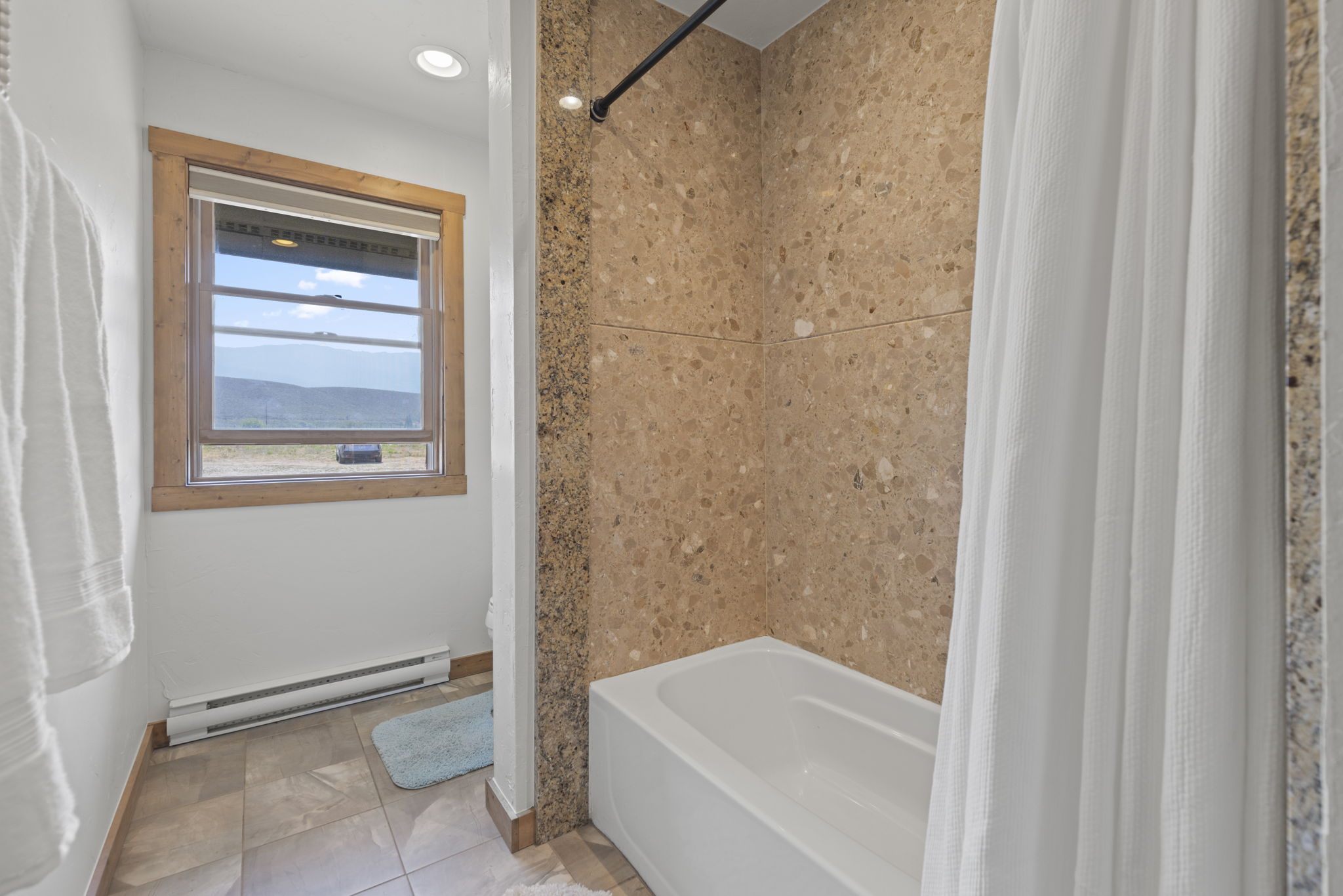 This bathroom features a white bathtub paired with a shower area enclosed by a textured, speckled stone-look surround. A window with light wood trim provides natural light and a view of the outdoors, while a white baseboard heater runs along the wall. The space is finished with neutral-toned floor tiles and a simple shower curtain, creating a functional and modest aesthetic.