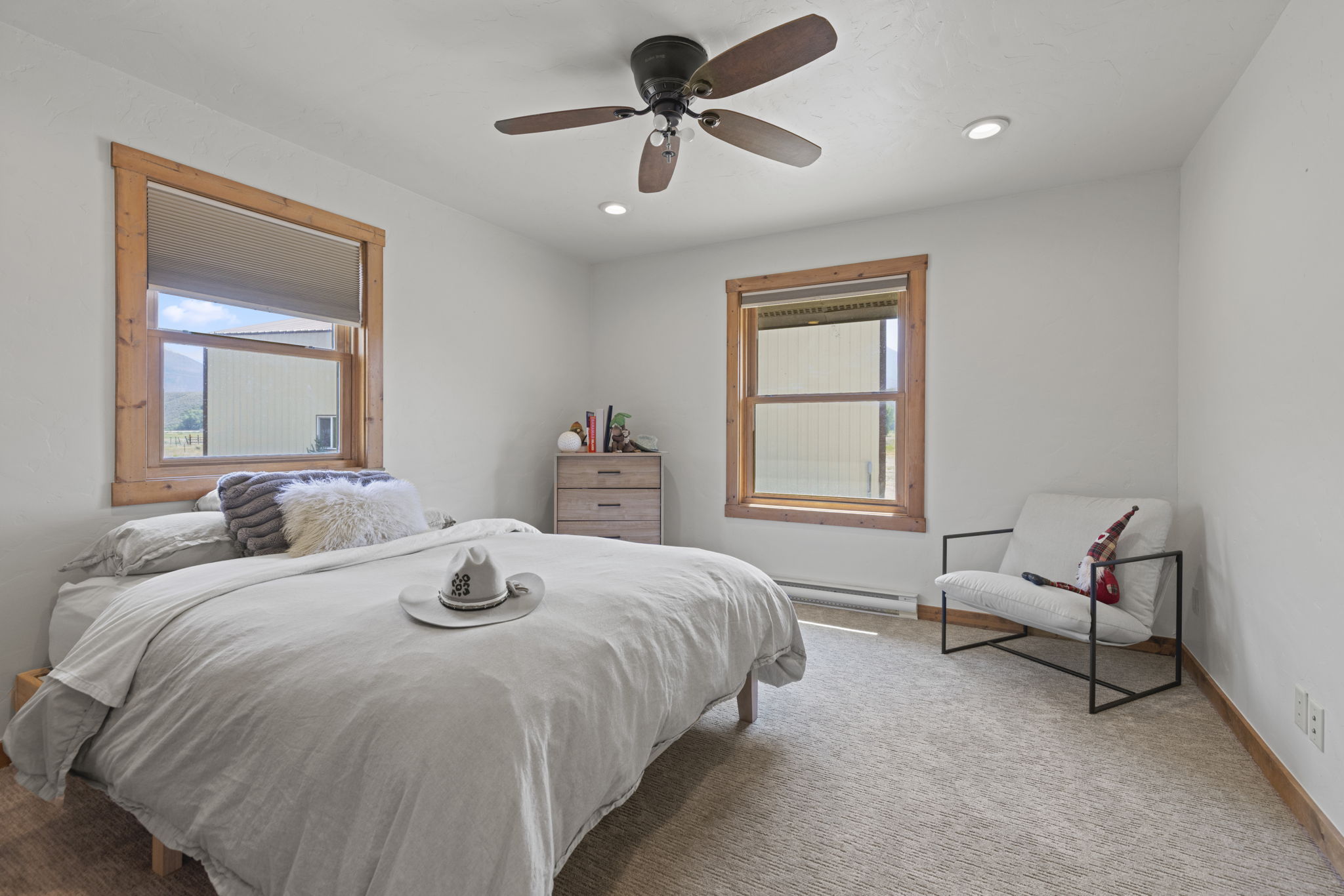 This bright and neutral bedroom features a queen-sized bed with a light-colored duvet and a decorative cowboy hat resting on top. The room is accented by two windows framed in natural wood trim, a light wood dresser, and a modern white armchair with a black metal frame. The space is finished with neutral carpeting and a ceiling fan, creating a clean and inviting atmosphere.