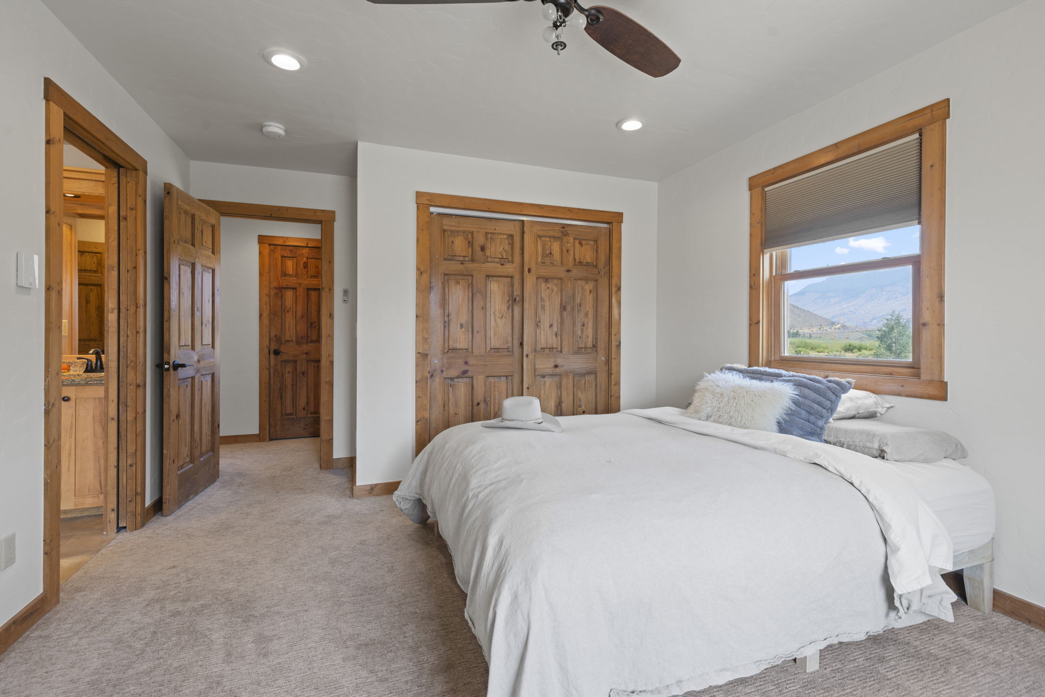 This cozy bedroom features a neutral color palette with light-toned walls and plush carpeting. A bed with a white duvet and decorative pillows is positioned near a window that offers a scenic view of the surrounding landscape. The room includes rustic wood-trimmed doors and a matching closet, creating a warm and inviting atmosphere.