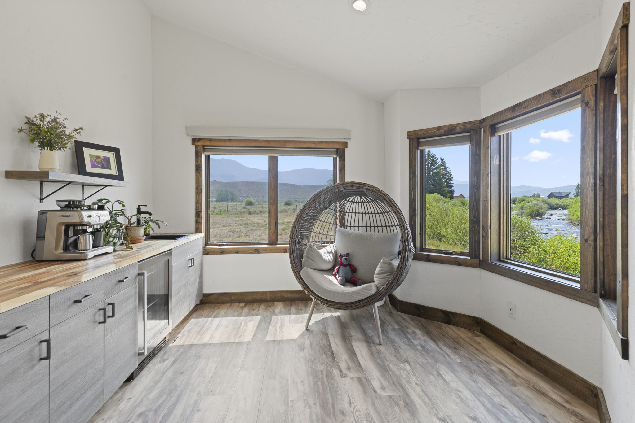 This bright and airy kitchenette area features light wood-toned cabinetry with a butcher block countertop and a built-in beverage refrigerator. A cozy, round wicker hanging chair sits in the corner, positioned to take advantage of the expansive windows that offer scenic views of the surrounding landscape and river. The space is accented by rustic wood window trim and a floating shelf, creating a warm and inviting atmosphere.