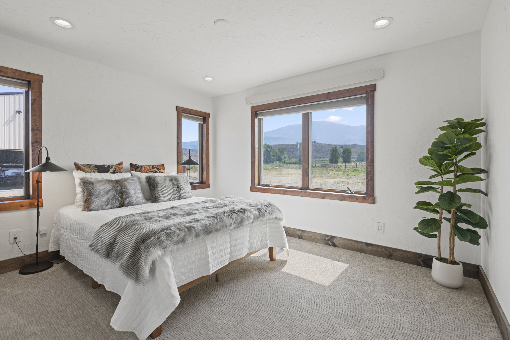 This bright and airy bedroom features a neutral color palette with warm wood trim around the windows and baseboards. The room is furnished with a bed dressed in white linens and a textured faux-fur throw, complemented by a potted plant in the corner and a sleek floor lamp. Large windows provide ample natural light and offer a scenic view of the surrounding landscape and mountains.