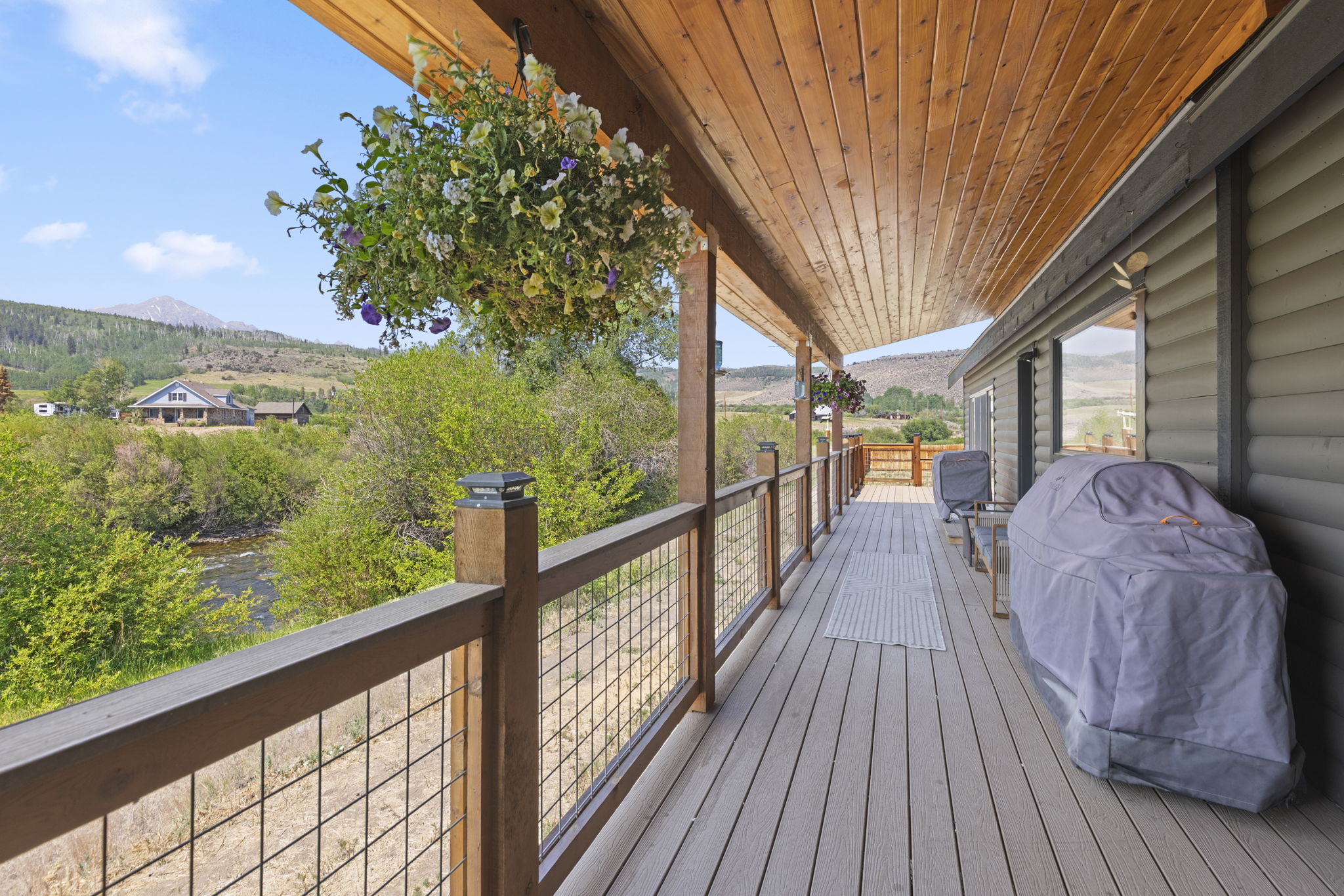 This expansive, covered wooden deck offers a serene outdoor living space with a rustic aesthetic, featuring a wire-mesh railing and a warm wood-paneled ceiling. A large, vibrant hanging flower basket adds a touch of color, while a covered grill and outdoor seating area suggest a perfect spot for relaxation. The deck overlooks a lush landscape with a flowing stream and distant mountains, providing a picturesque and tranquil setting.