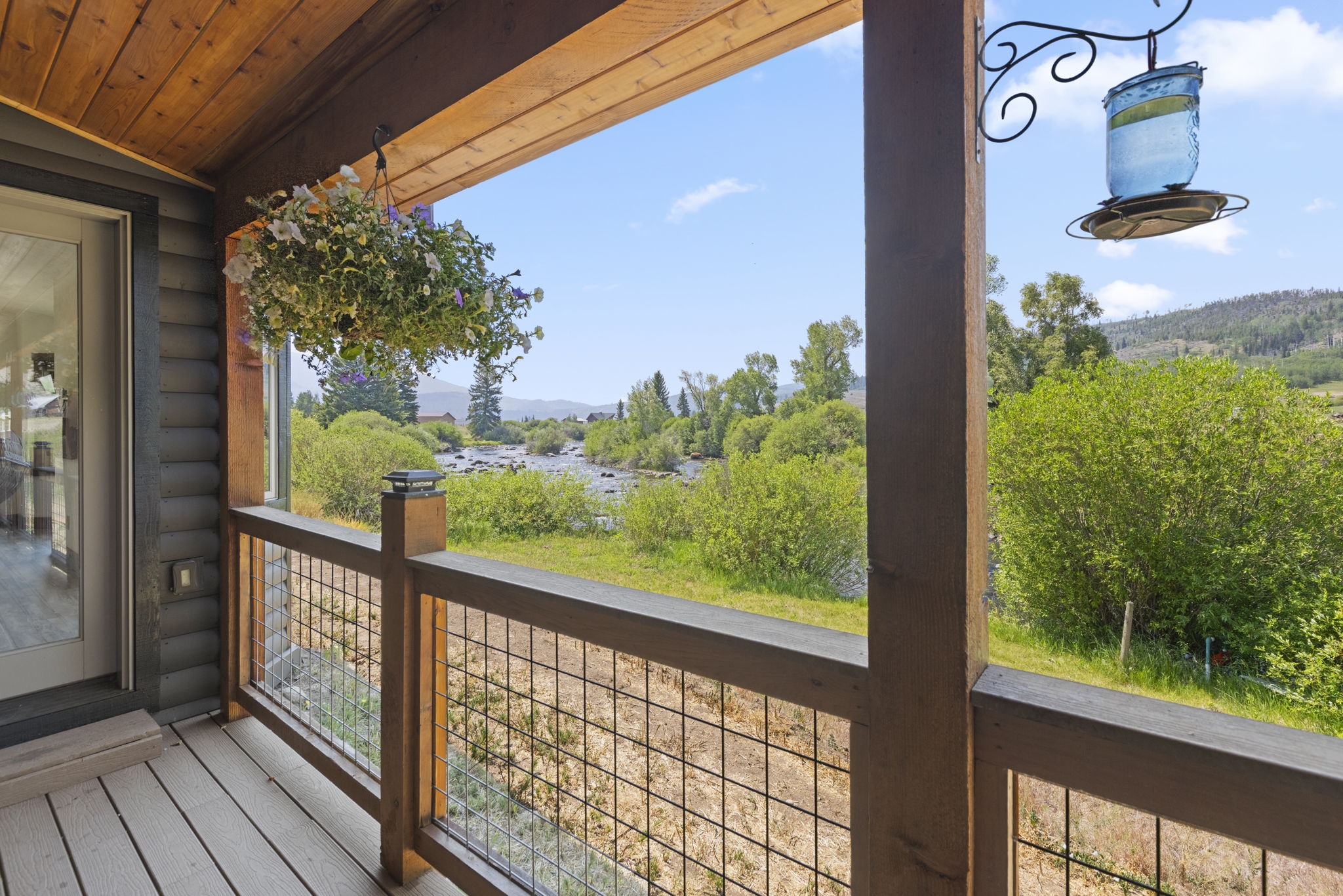 This inviting wooden deck offers a serene, elevated perspective overlooking a lush river landscape and distant mountains. The space features a rustic railing with wire mesh panels, a hanging flower basket, and a bird feeder, creating a peaceful outdoor retreat. The view captures the natural beauty of the surrounding greenery and flowing water, emphasizing a tranquil connection to the outdoors.
