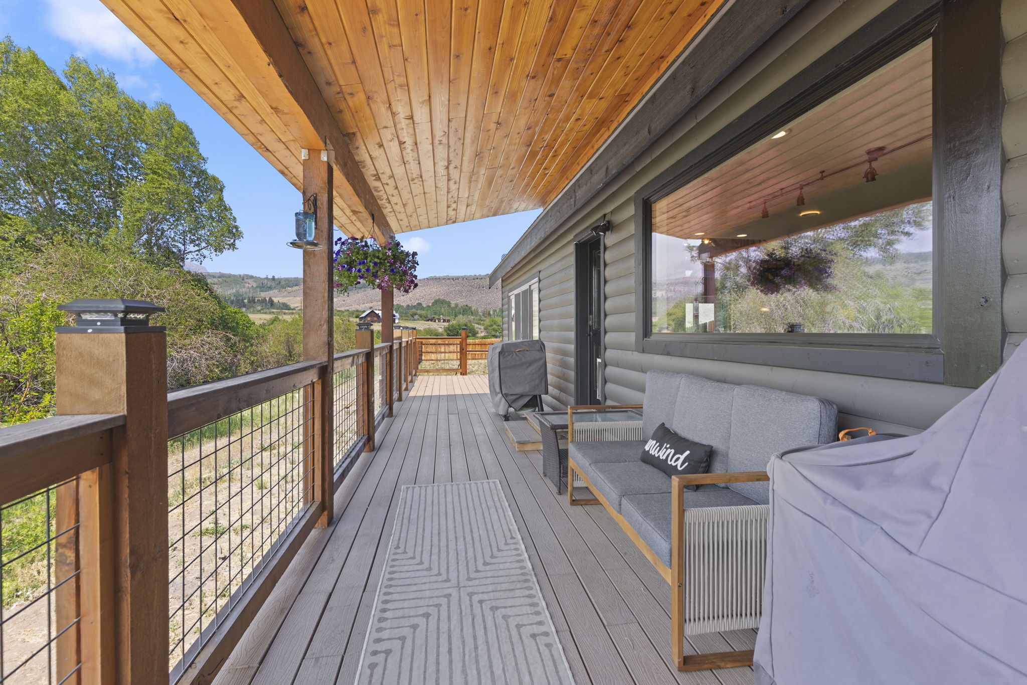 This spacious, covered wooden deck offers a serene outdoor living space with a rustic log-style exterior wall and a large window reflecting the surrounding landscape. The deck is furnished with a comfortable gray sofa featuring an 'unwind' throw pillow and is accented by a patterned area rug and hanging flower baskets. The perspective looks down the length of the deck, highlighting the wire-railing fence that provides an unobstructed view of the rolling hills and natural scenery beyond.