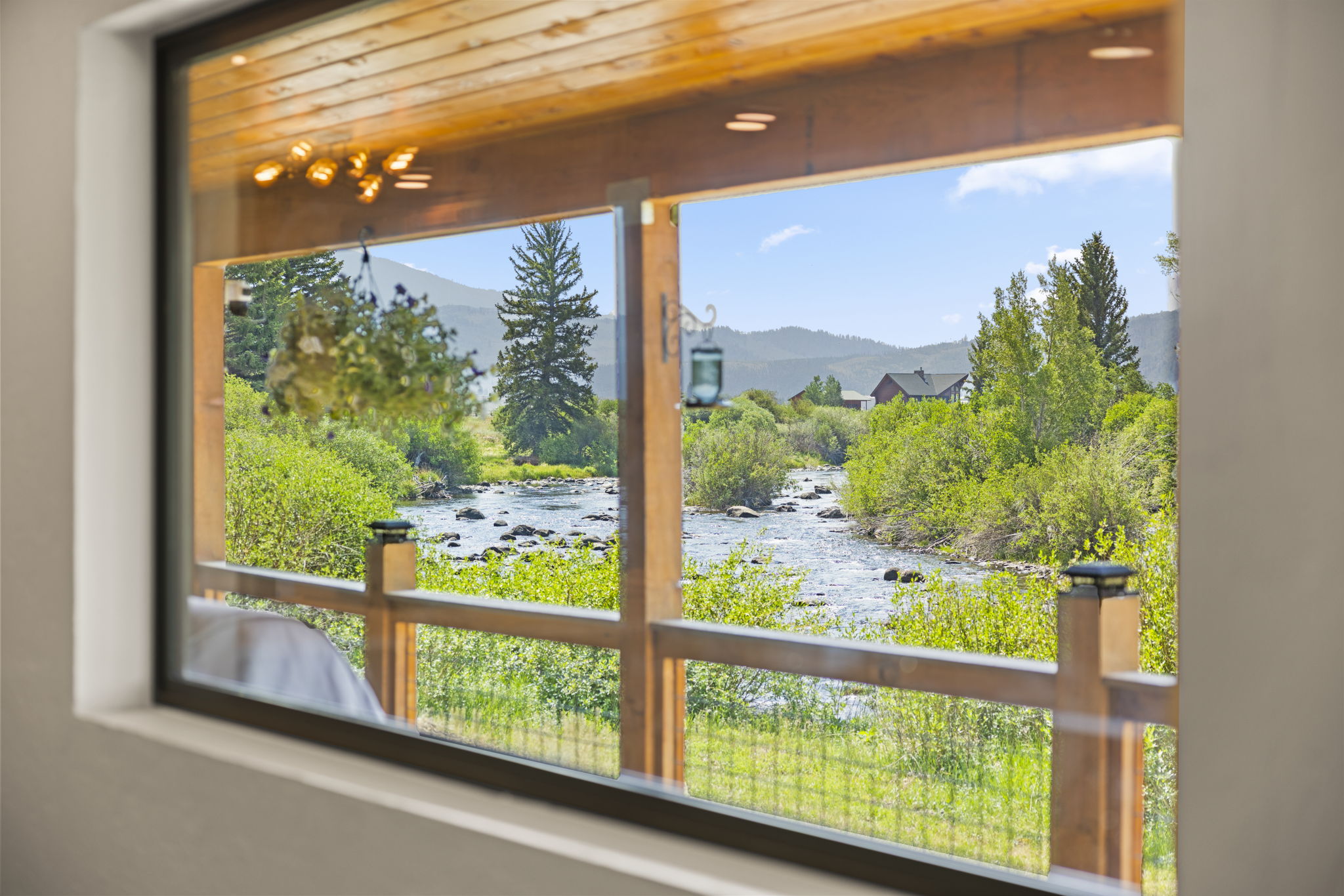This interior perspective looks out through a large, expansive window that frames a picturesque view of a rushing river and lush green landscape. The foreground features a wooden deck railing and a hanging plant, creating a seamless transition between the indoor living space and the natural scenery. The warm wood-paneled ceiling and soft interior lighting enhance the cozy, inviting atmosphere of this scenic mountain retreat.