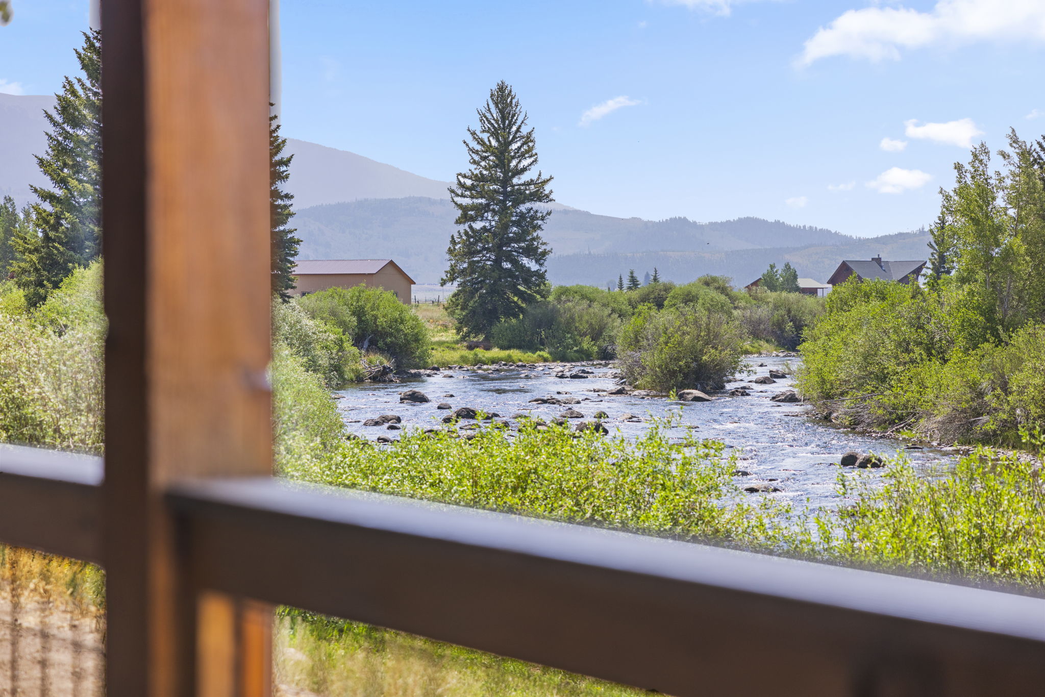 This view is captured from a wooden deck or balcony, looking out over a scenic river landscape. The foreground features a wooden railing that frames the natural vista, which includes a flowing stream, lush green vegetation, and a prominent evergreen tree. In the distance, rolling mountains under a bright blue sky create a serene and picturesque setting.
