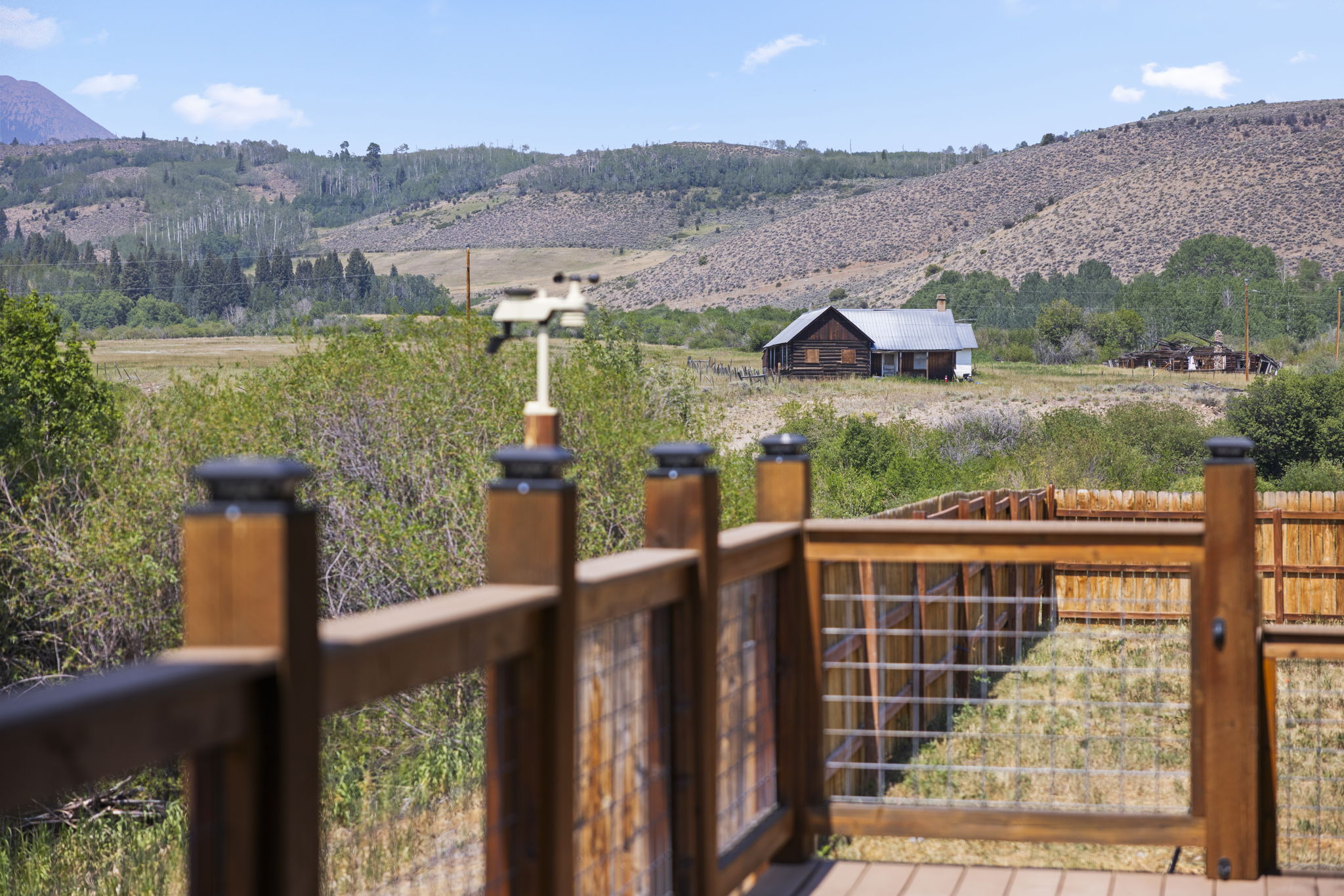 This perspective is captured from a wooden deck, showcasing a rustic railing with wire mesh panels that frame a scenic rural landscape. In the distance, a weathered wooden cabin sits amidst a sprawling field, set against a backdrop of rolling hills and a clear blue sky. The scene evokes a sense of peaceful, expansive country living with a focus on the natural surroundings.