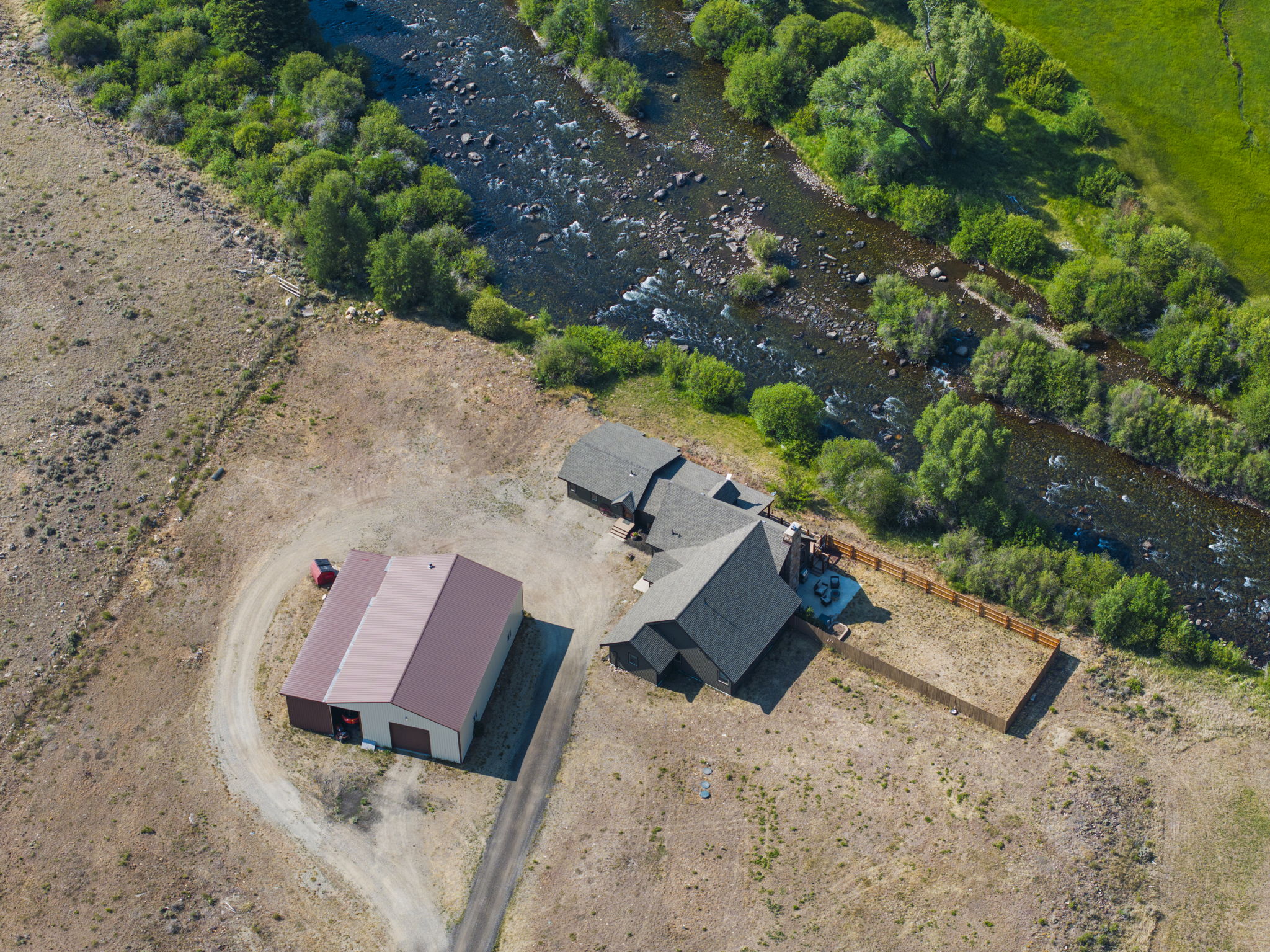 This high-angle aerial perspective captures a rural property nestled beside a winding, rocky river. The site features a primary residence with a dark-toned roof, an adjacent detached metal-roofed workshop or garage, and a fenced-in yard area. The surrounding landscape is characterized by open, dry terrain transitioning into lush, tree-lined riverbanks, offering a serene and secluded atmosphere.