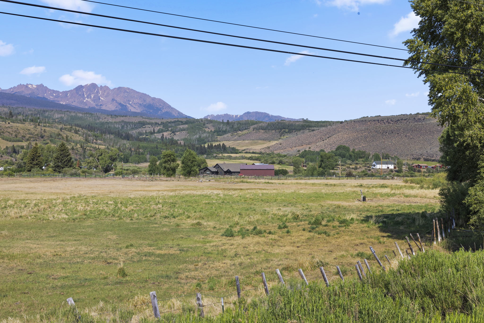 This wide-angle landscape shot captures a sprawling rural property set against a dramatic backdrop of rugged, snow-dusted mountain peaks. In the foreground, a wire fence borders a vast, sun-drenched meadow, while a cluster of residential buildings with dark siding and a prominent red-roofed structure sits nestled in the valley. The scene conveys a sense of expansive, peaceful mountain living with clear blue skies and lush, natural surroundings.