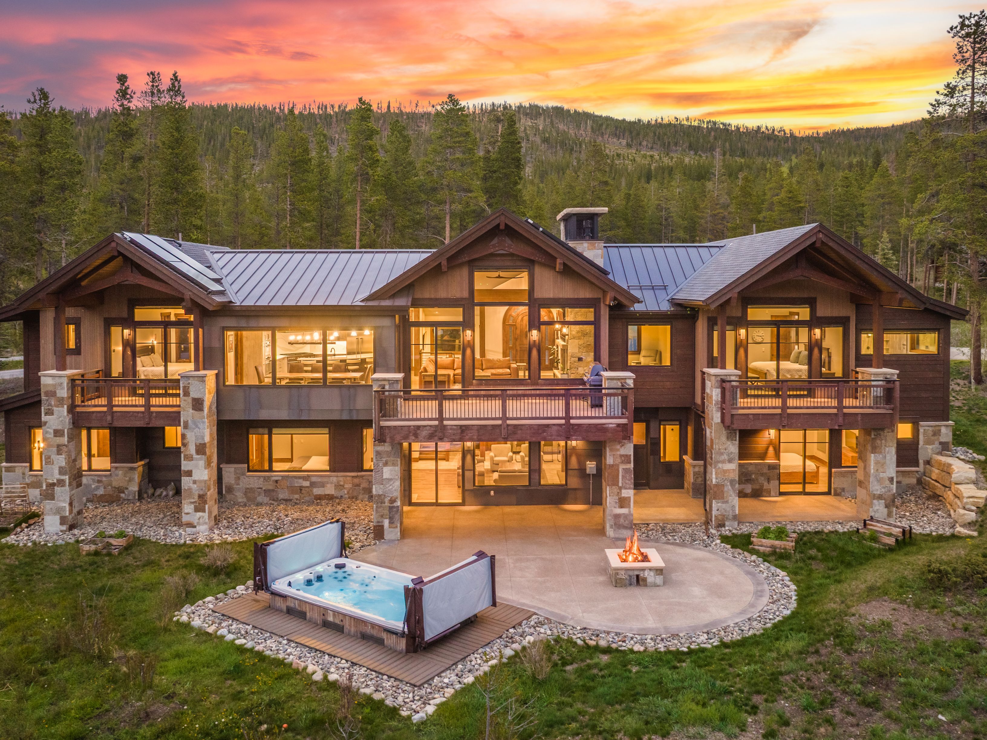 This stunning mountain estate features a multi-level design with a blend of dark wood siding and natural stone accents. The rear facade showcases expansive windows, multiple balconies, and a spacious patio complete with a fire pit and a hot tub, all set against a backdrop of a lush forest at sunset. The cinematic perspective captures the home's grand scale and seamless integration with the surrounding wilderness.