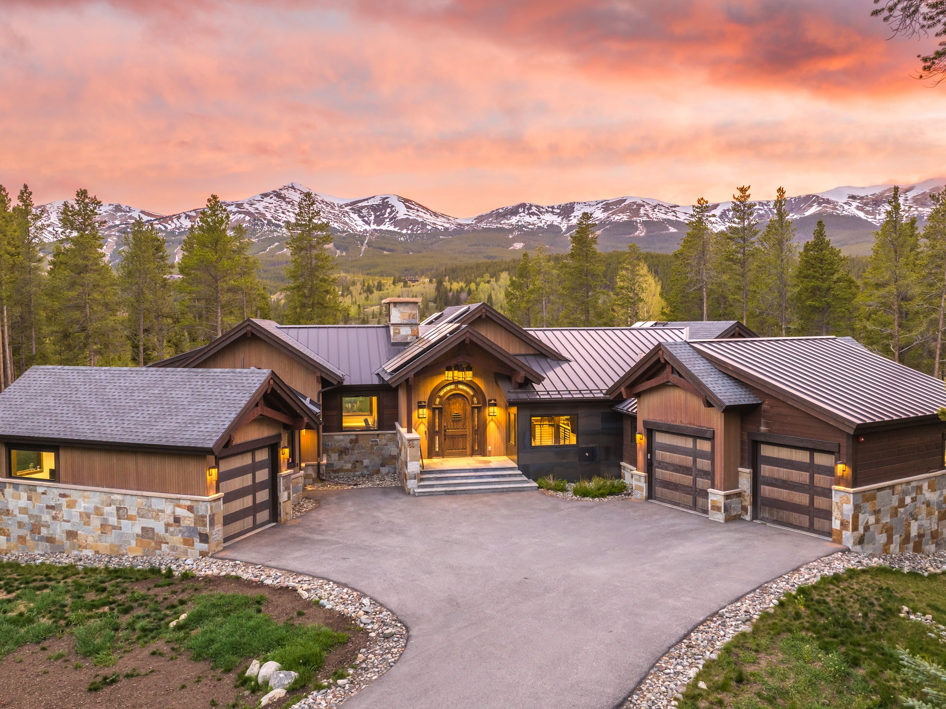 This stunning mountain estate features a rustic-modern architectural style with a blend of dark wood siding, stone accents, and a metal roof. The front facade showcases a grand arched entryway, three garage bays, and a paved driveway that curves through the landscaped grounds. Set against a backdrop of snow-capped mountains and a vibrant sunset, the home exudes a luxurious and serene atmosphere.