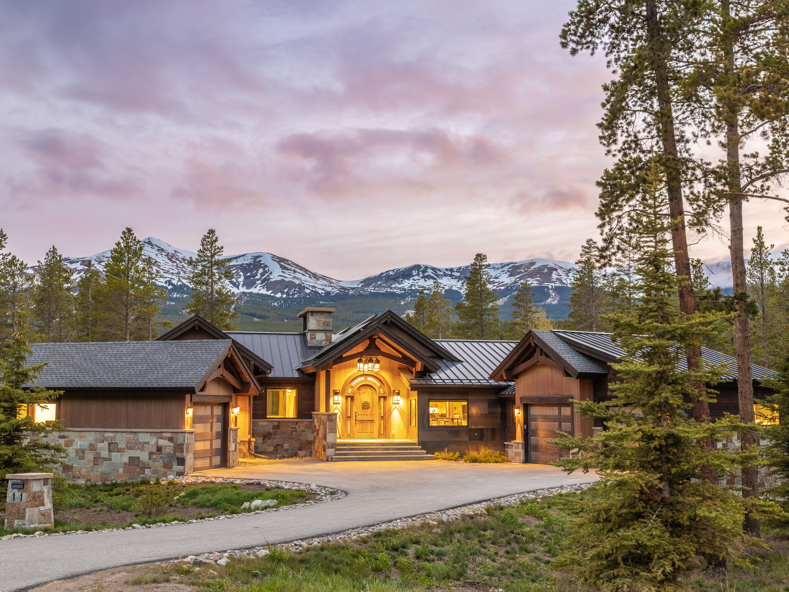 This stunning mountain residence features a rustic-modern aesthetic with a blend of dark wood siding, stone accents, and a metal roof. The symmetrical design highlights a grand, arched wooden front door illuminated by warm lighting, flanked by two garage bays and set against a backdrop of snow-capped peaks at dusk. A paved driveway leads up to the entrance, framed by mature pine trees and natural landscaping, creating an inviting and luxurious alpine retreat.