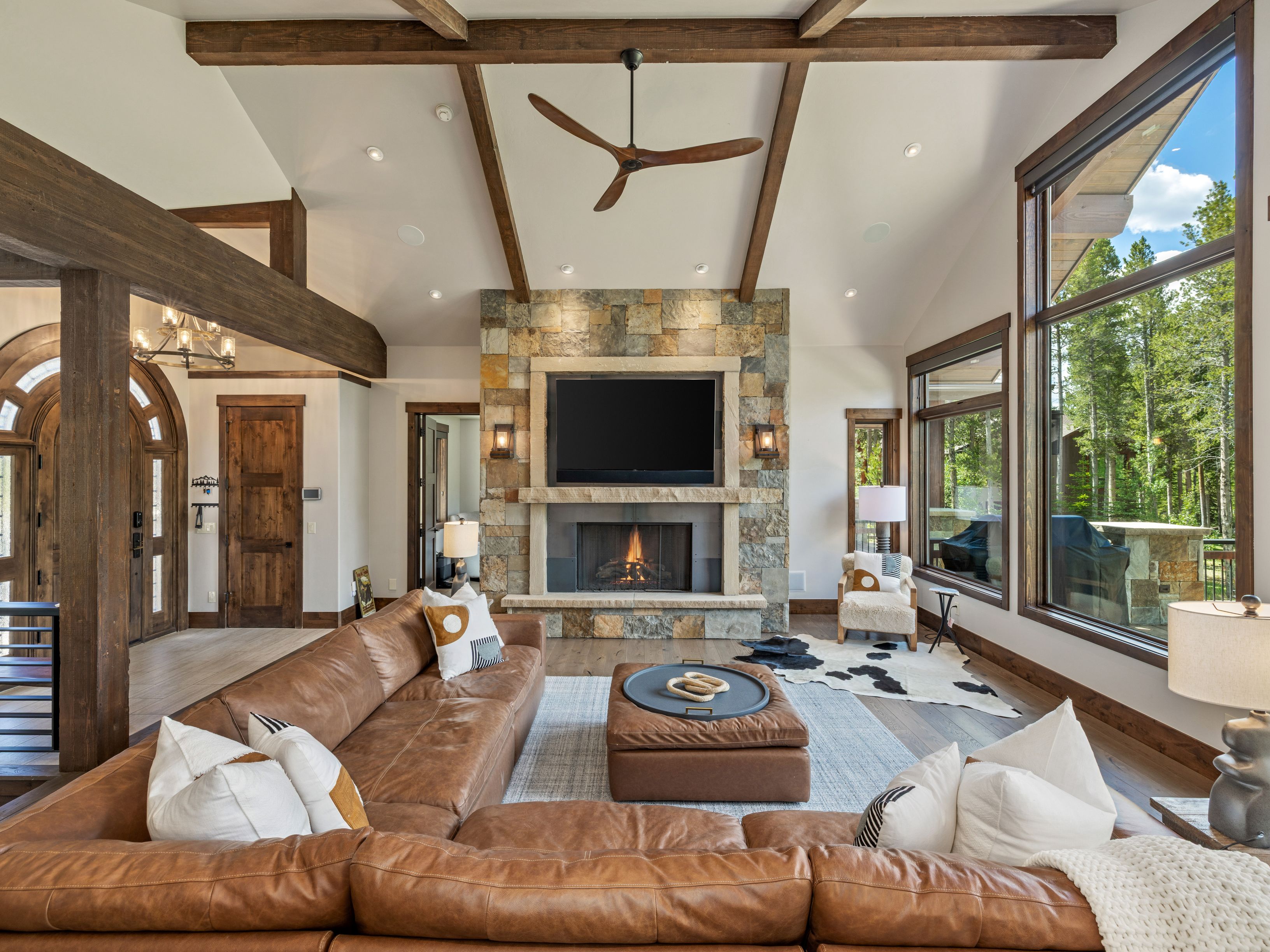 This spacious living room features a vaulted ceiling with exposed wooden beams and a prominent stone-clad chimney breast housing a television and fireplace. A large, plush leather sectional sofa provides ample seating, centered around a matching ottoman on a textured area rug. Large windows offer expansive views of the surrounding forest, creating a bright and inviting atmosphere with a rustic, mountain-lodge aesthetic.