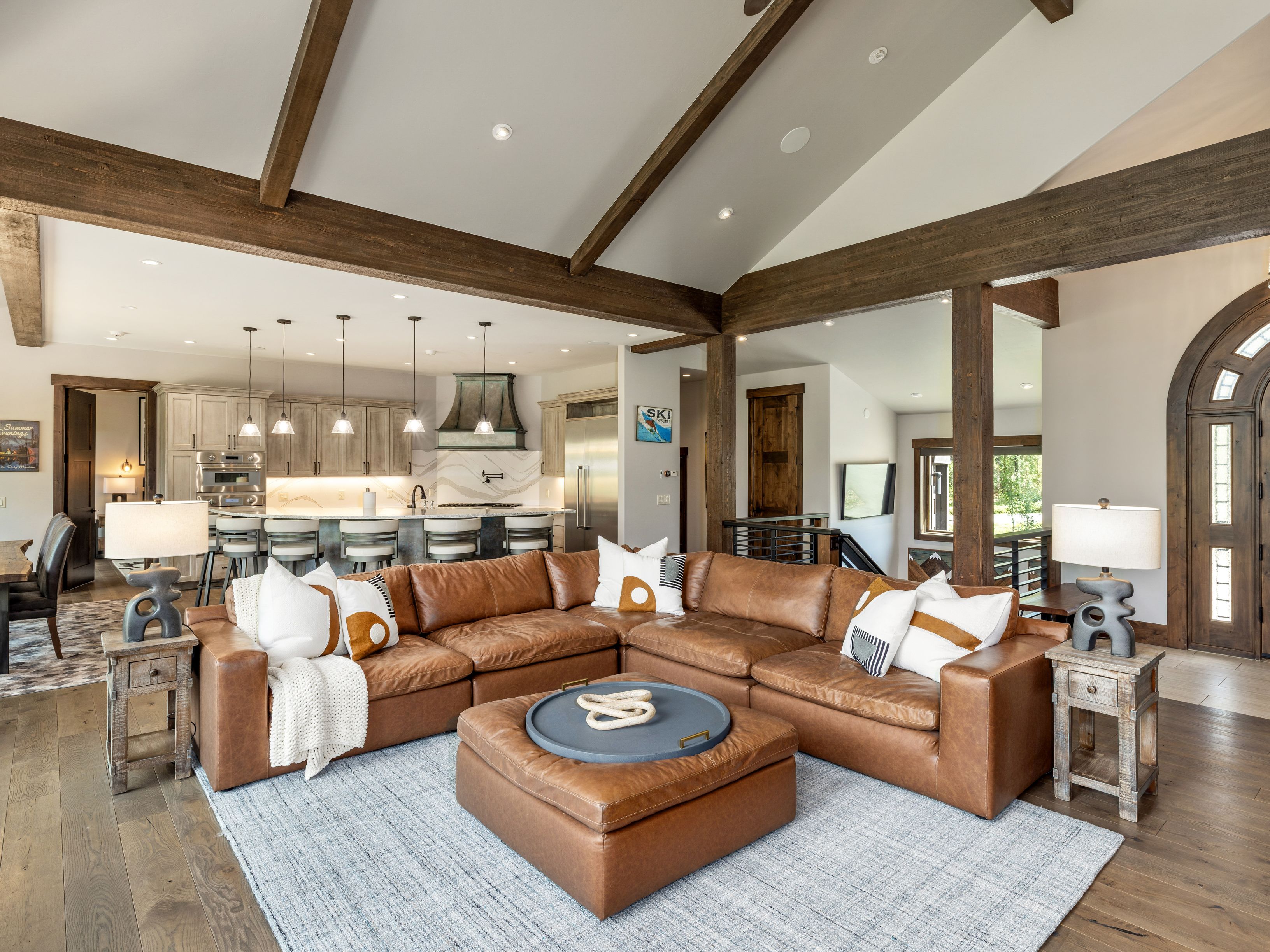This spacious open-concept living area features a large, rich brown leather sectional sofa centered on a light-colored area rug, creating a warm and inviting atmosphere. The room is defined by vaulted ceilings with exposed dark wood beams and flows seamlessly into a well-appointed kitchen with a breakfast bar. The aesthetic blends rustic charm with modern comfort, highlighted by decorative lamps on side tables and a large ottoman serving as a central coffee table.