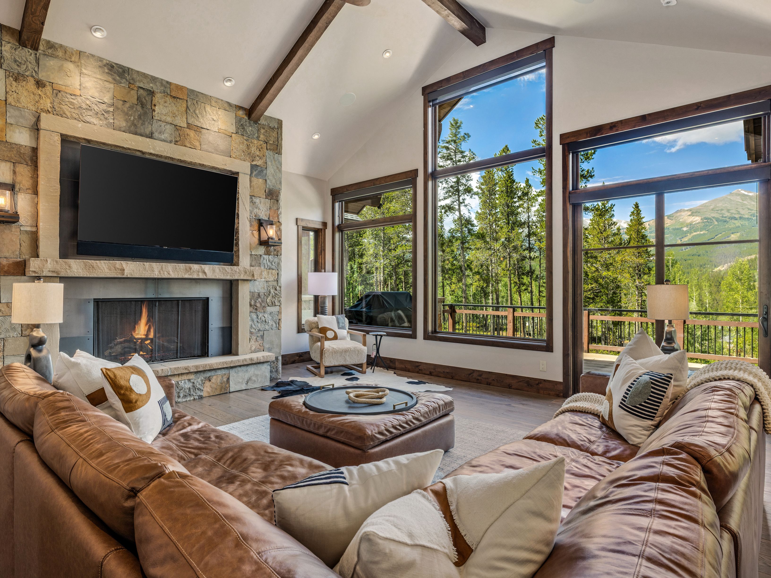 This spacious living room features a vaulted ceiling with exposed wooden beams and a prominent stone-clad chimney breast housing a fireplace and a large television. A plush, L-shaped leather sectional provides comfortable seating, centered around a large leather ottoman. Tall windows frame expansive views of the surrounding forest and mountains, filling the room with natural light and creating a warm, mountain-lodge aesthetic.