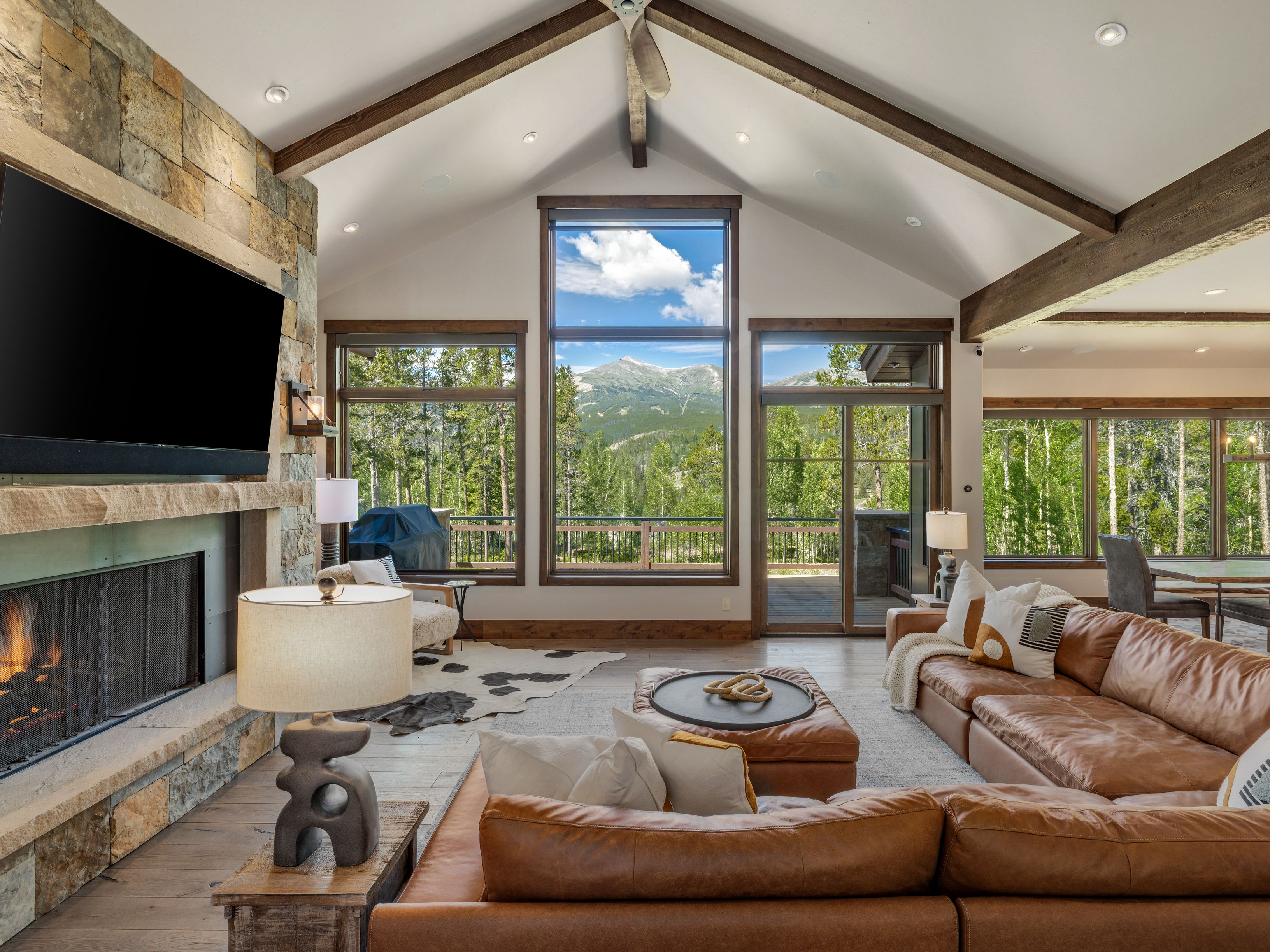 This spacious living room features a vaulted ceiling with exposed timber beams and a prominent stone-clad chimney breast housing a fireplace. A large leather sectional sofa faces the fireplace, while expansive windows provide a panoramic view of the surrounding forest and distant mountains. The room is decorated with a cowhide rug, a leather ottoman, and warm lighting, creating a cozy and inviting mountain-lodge atmosphere.