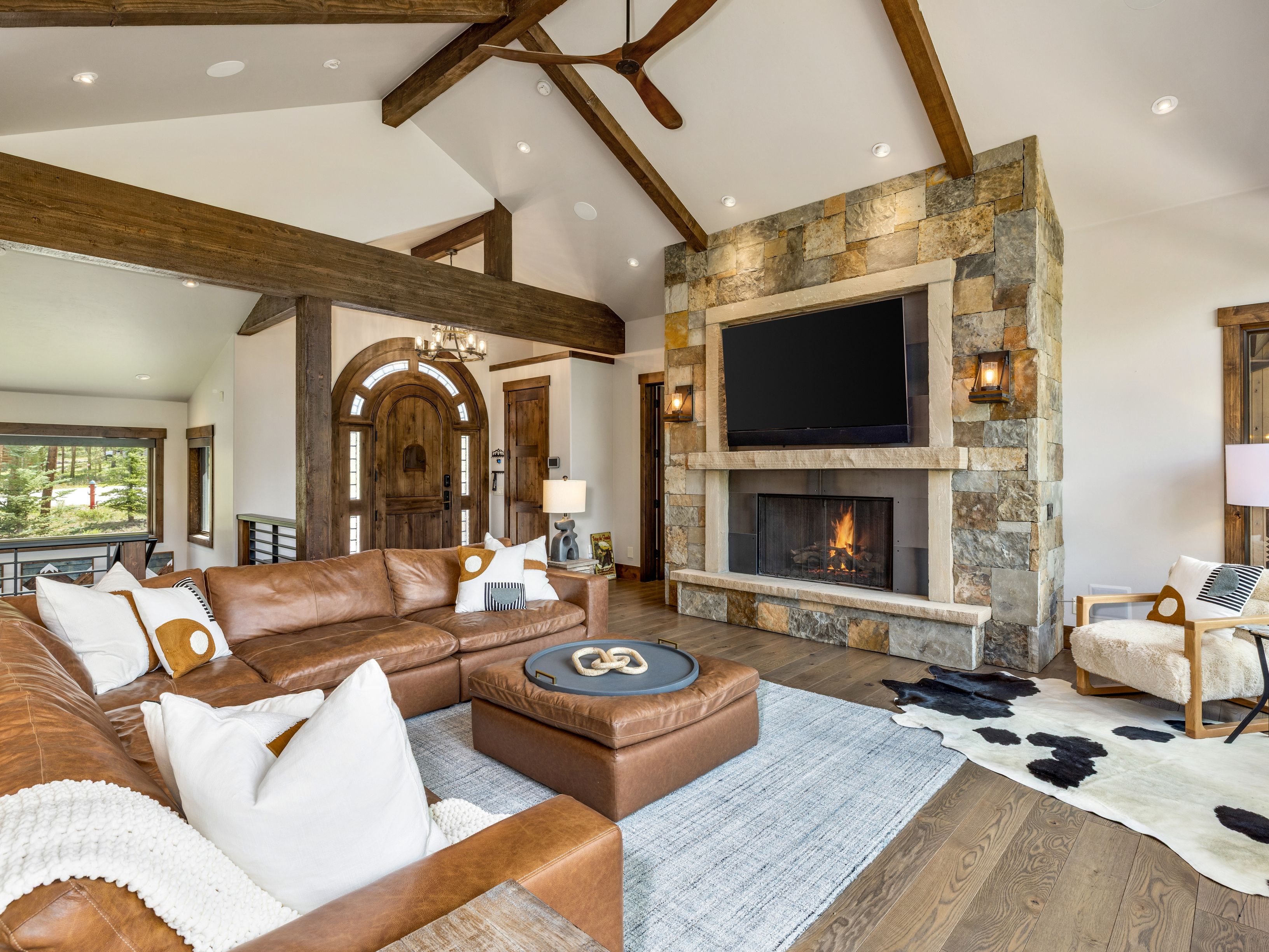 This spacious living room features a vaulted ceiling with exposed wooden beams and a prominent stone-clad chimney breast housing a fireplace and television. A large leather sectional sofa and matching ottoman are arranged on a textured area rug, complemented by a cowhide rug and a cozy armchair. The open-concept layout provides a clear view of the arched wooden front door, creating a warm, rustic, and inviting atmosphere.