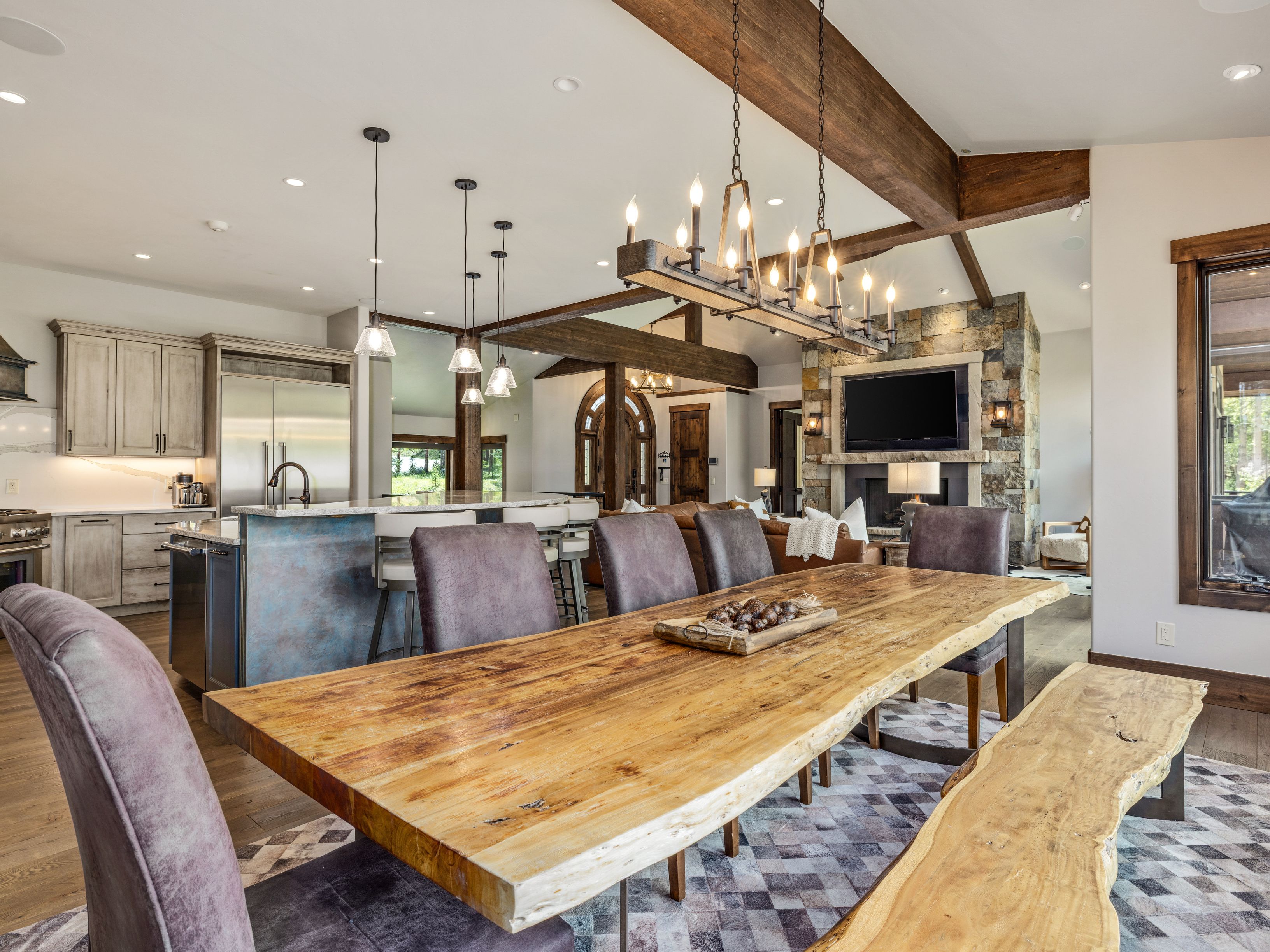 This open-concept dining area features a prominent live-edge wood dining table paired with a matching bench and upholstered chairs, set atop a geometric patterned rug. The space flows seamlessly into a kitchen with a blue-toned island and a living area anchored by a stone-clad chimney breast with an integrated television. Exposed wooden ceiling beams and warm pendant lighting enhance the rustic, high-end aesthetic of this expansive great room.