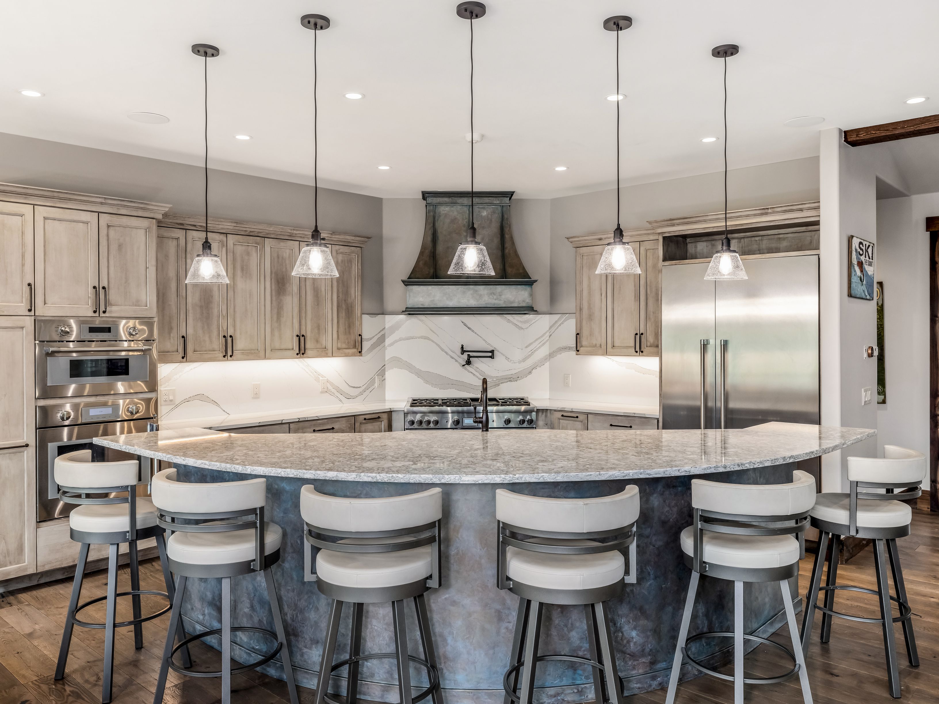 This spacious kitchen features a large, curved island with a stone countertop and five bar stools, serving as the central focal point. The space is equipped with stainless steel appliances, including a double wall oven and a large refrigerator, complemented by light-toned cabinetry and a decorative range hood. Five pendant lights hang above the island, providing warm illumination against the neutral-toned backsplash and wood flooring.