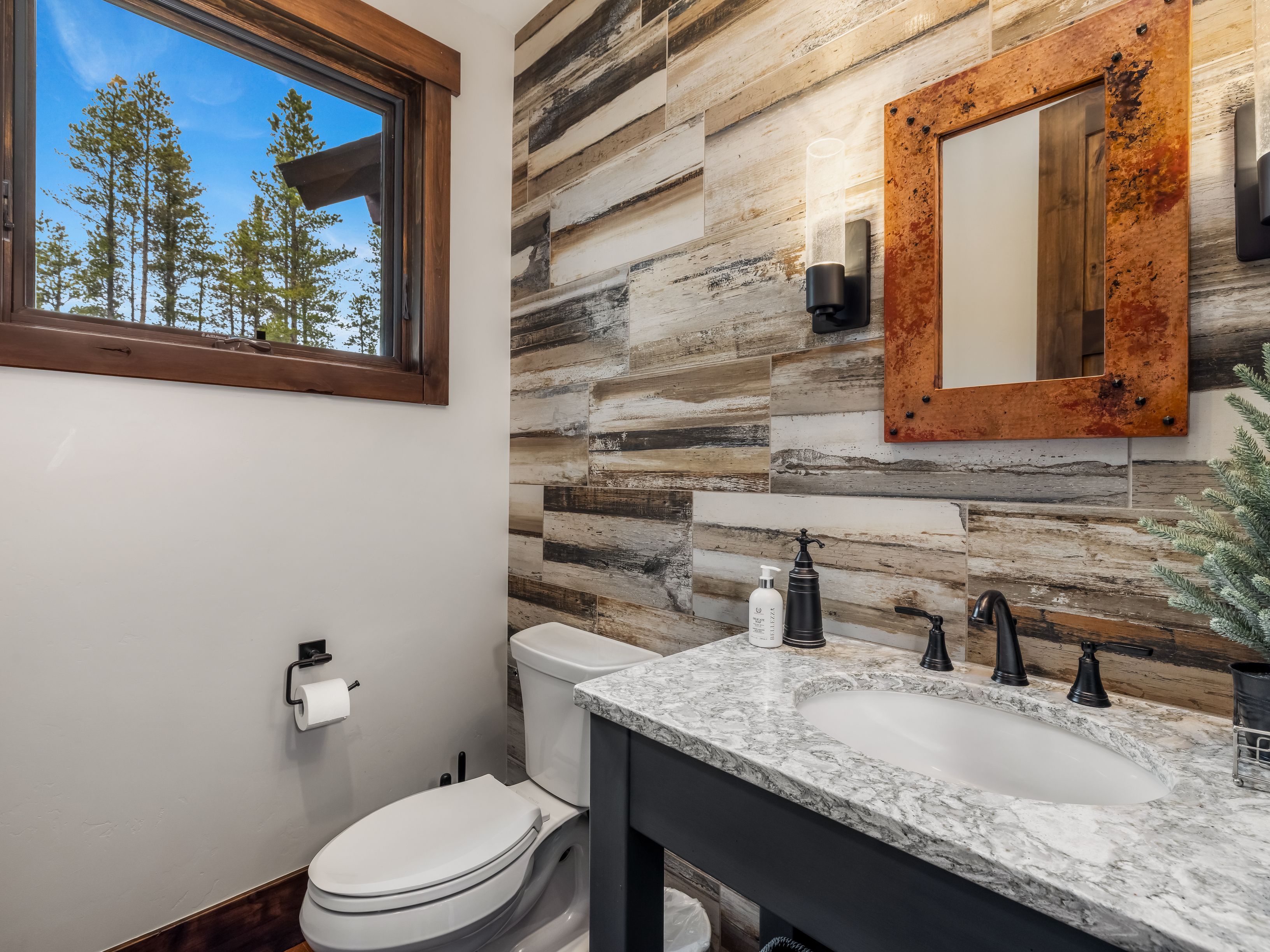 This rustic-style powder room features a distinctive wall clad in horizontal, multi-toned wood-look tiles that provide a textured backdrop. A vanity with a stone-patterned countertop and a dark base sits beneath a unique, weathered metal-framed mirror, while a window above the toilet offers a view of the surrounding forest. The space is accented by matte black fixtures and warm wall lighting, creating a cozy and earthy atmosphere.