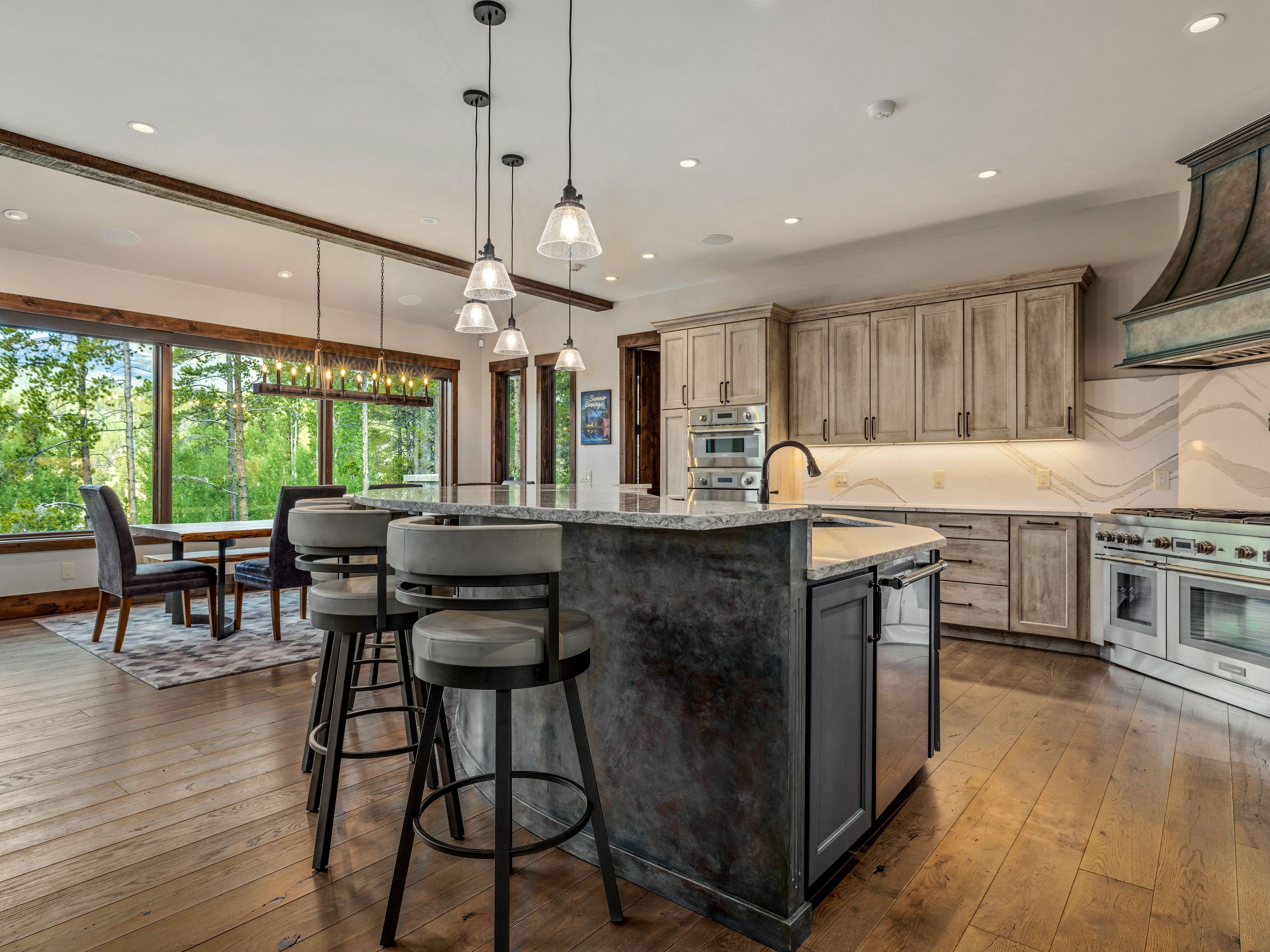 This spacious, open-concept kitchen features a large central island with bar seating and a dark, textured base, complemented by light-toned cabinetry and stone countertops. A dining area is positioned near a wall of large windows that provide abundant natural light and views of the surrounding forest. The space is accented by rustic wooden ceiling beams and modern pendant lighting, creating a warm and inviting atmosphere.