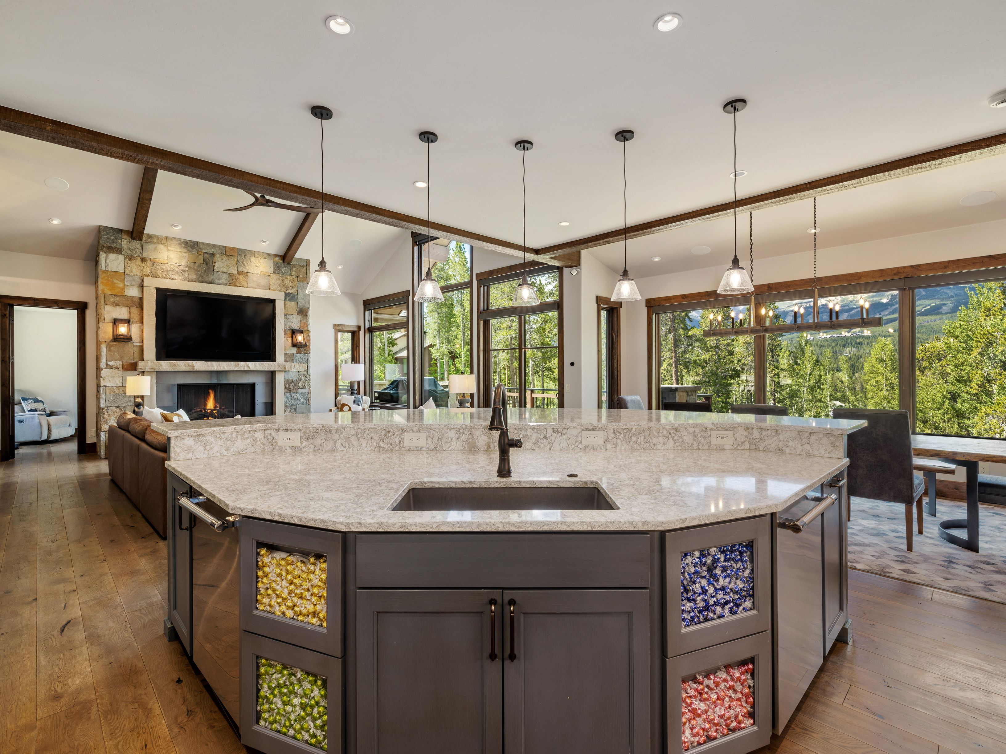 This spacious, open-concept kitchen features a large, multi-sided island with a stone countertop and a central sink, serving as the focal point of the room. The island base is uniquely designed with glass-fronted cabinets displaying colorful wrapped candies, adding a playful touch to the sophisticated, rustic-modern aesthetic. Beyond the kitchen, the living area showcases a stone-clad chimney breast and large windows that frame a scenic mountain view, creating a seamless connection between the interior and the natural landscape.