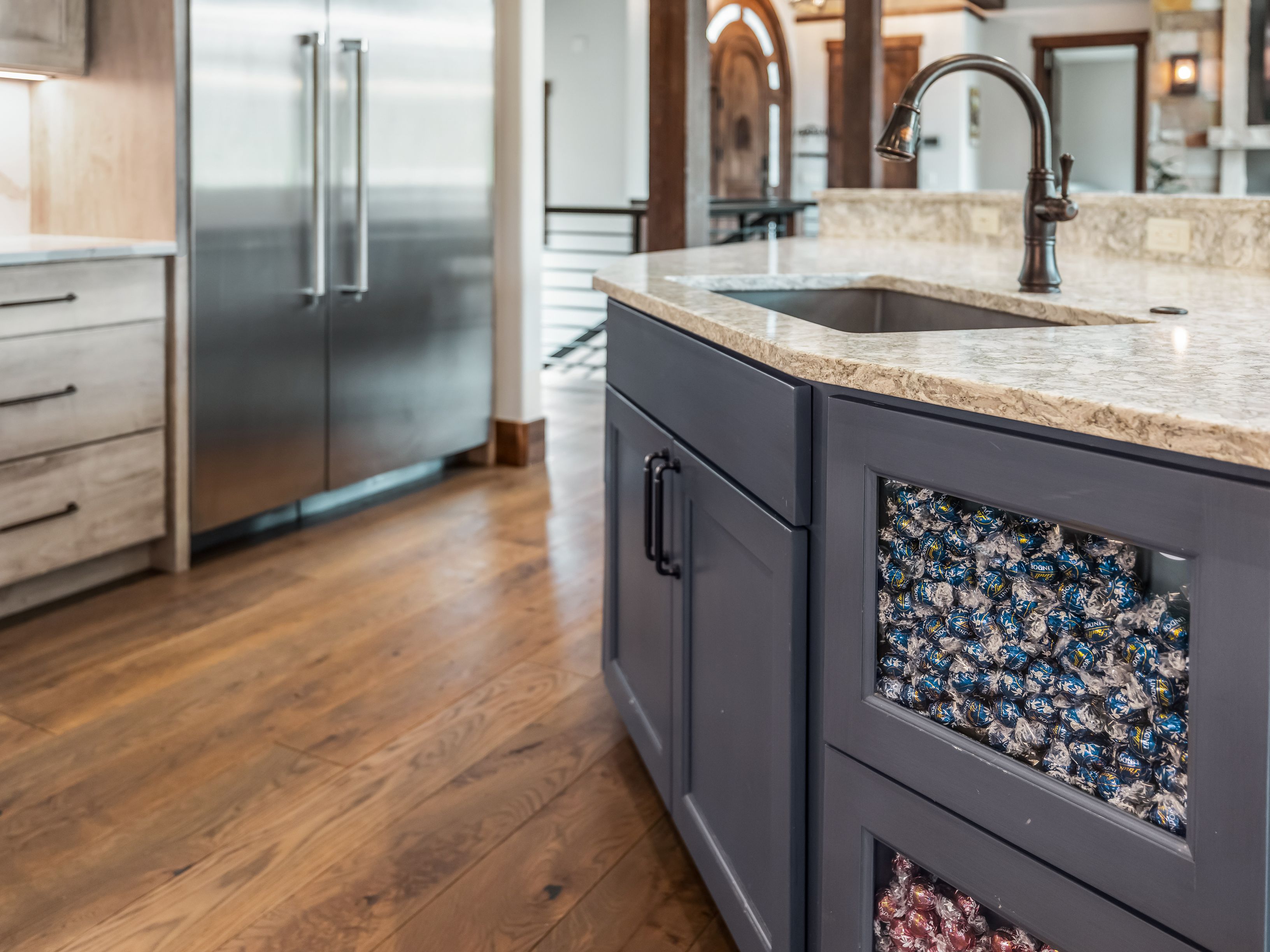 This kitchen features a dark-toned island with unique glass-fronted drawers filled with colorful wrapped chocolates, serving as a playful focal point. The space includes a stainless steel refrigerator, light wood cabinetry, and a stone countertop with an undermount sink and a dark metal faucet. The warm wood flooring and open layout create an inviting atmosphere, with a glimpse into the adjacent living area.