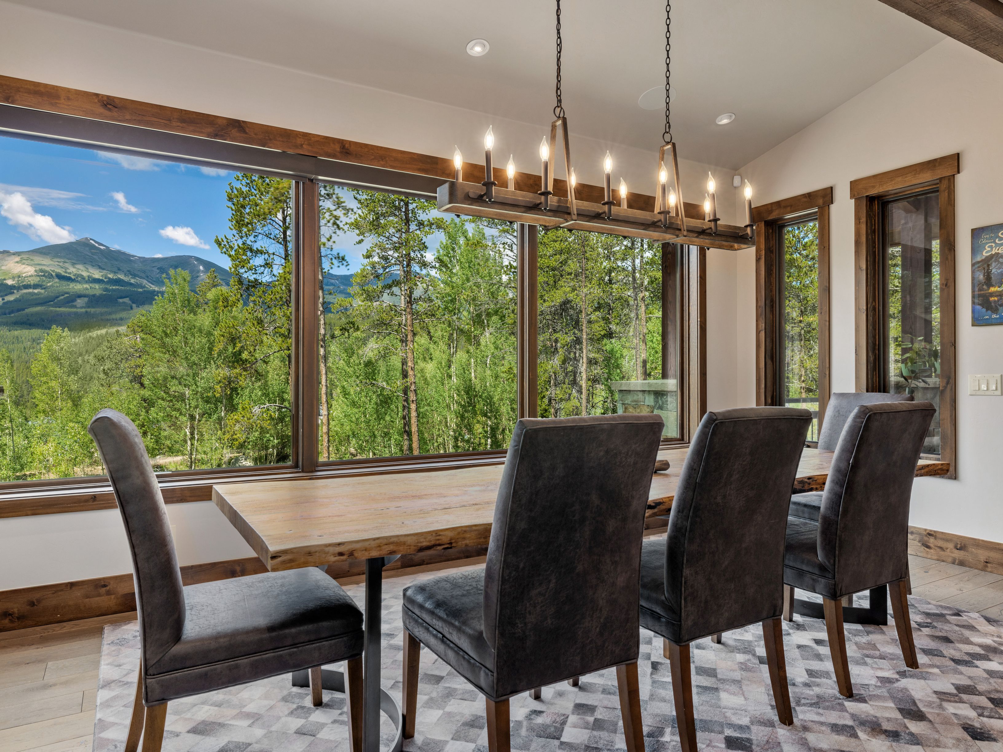 This dining room features a rustic live-edge wood table surrounded by four dark upholstered chairs, set atop a geometric patterned rug. A large, multi-light chandelier hangs above the table, while expansive windows provide a panoramic view of a lush forest and distant mountains. The space is framed by warm wood trim, creating a cozy, mountain-inspired atmosphere.