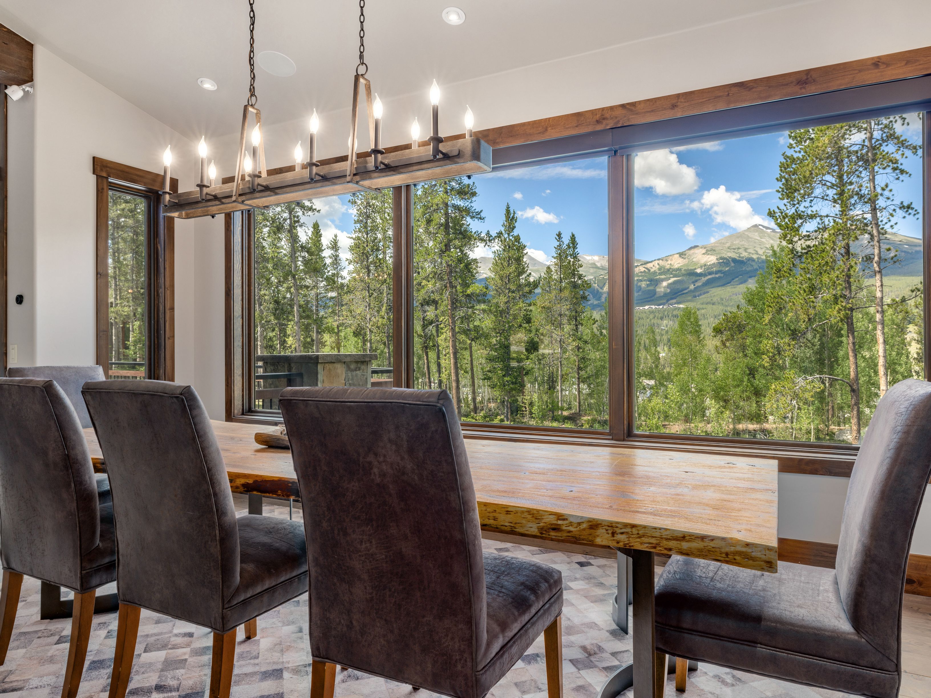 This dining room features a rustic live-edge wood table surrounded by four upholstered chairs, positioned to take full advantage of the expansive windows. A linear chandelier with multiple bulbs hangs above the table, providing warm lighting against the backdrop of a scenic mountain and forest view. The room is grounded by a patterned rug, creating a cozy and inviting atmosphere perfect for enjoying the natural surroundings.