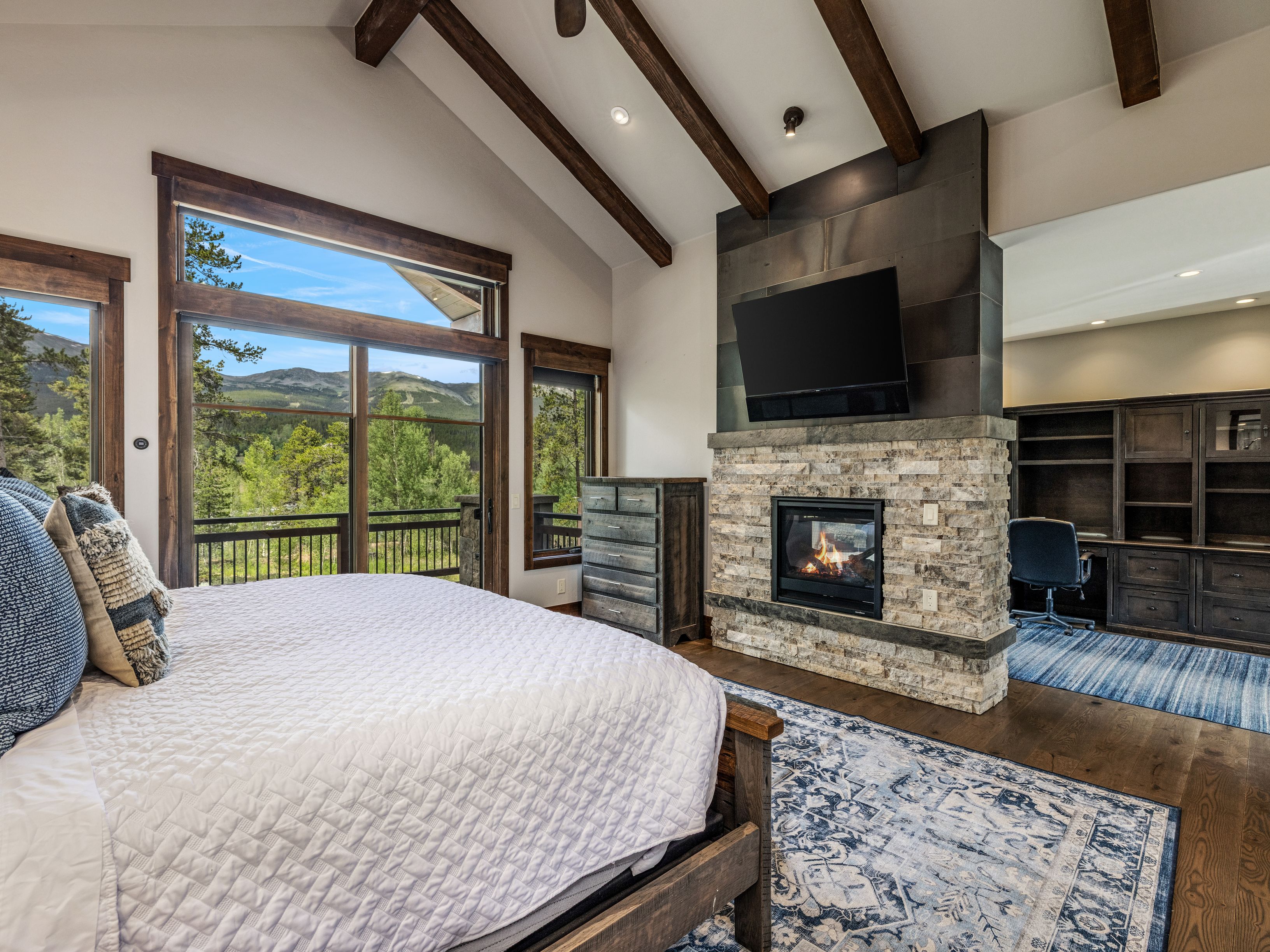 This primary bedroom features a vaulted ceiling with exposed wooden beams and a prominent stone-clad chimney breast that separates the sleeping area from an adjacent office space. A large bed with a textured blue quilt sits in the foreground, while expansive windows provide a scenic view of the surrounding mountains and forest. The room is anchored by a patterned area rug and includes a dark wood dresser, creating a cozy, rustic-inspired atmosphere.