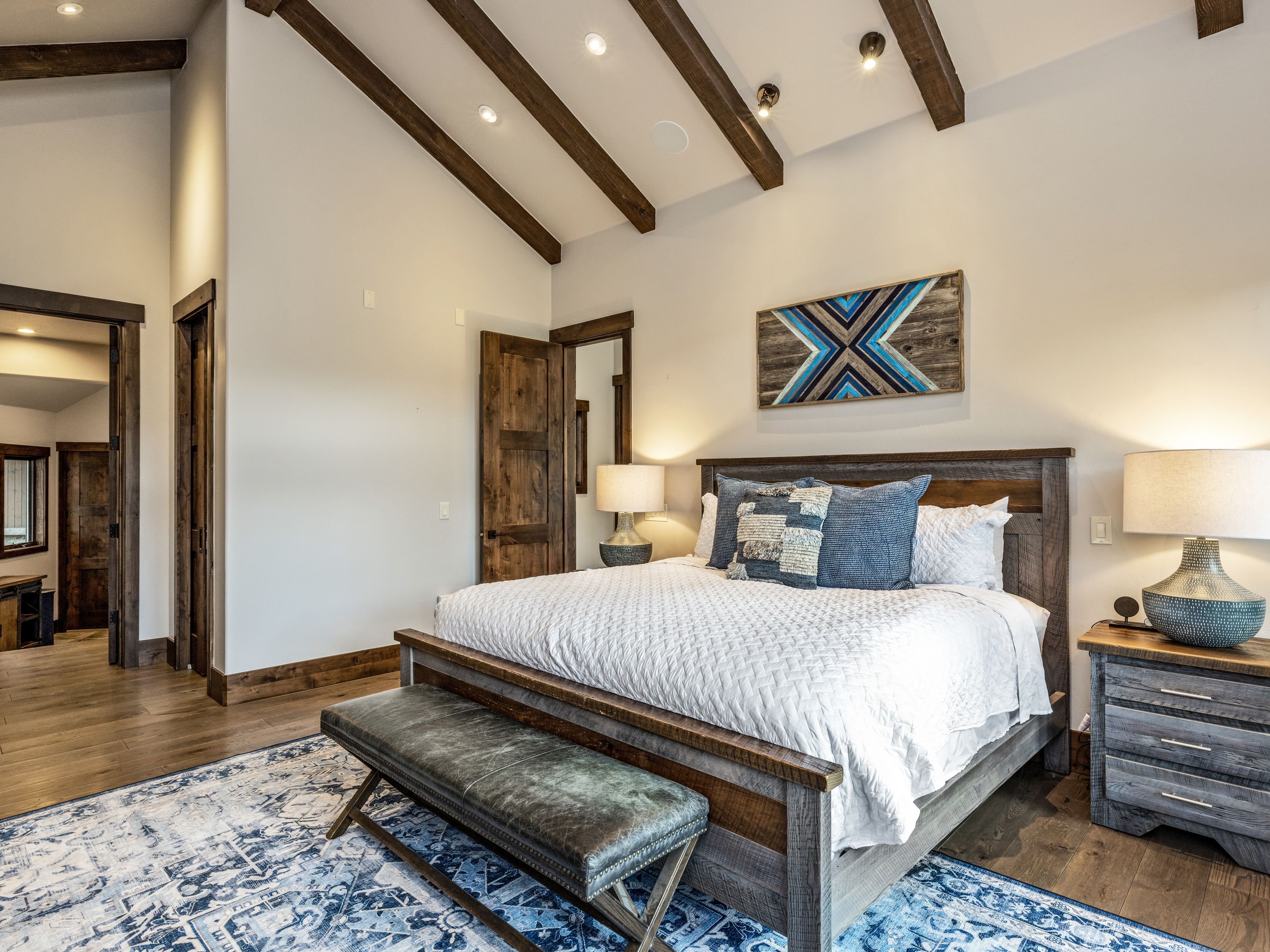 This primary bedroom features a rustic aesthetic with exposed wooden ceiling beams and a vaulted ceiling. A large bed with a dark wood frame is centered in the room, complemented by a patterned area rug and a leather bench at the foot of the bed. The space is illuminated by warm bedside lamps and recessed lighting, creating a cozy and inviting atmosphere.