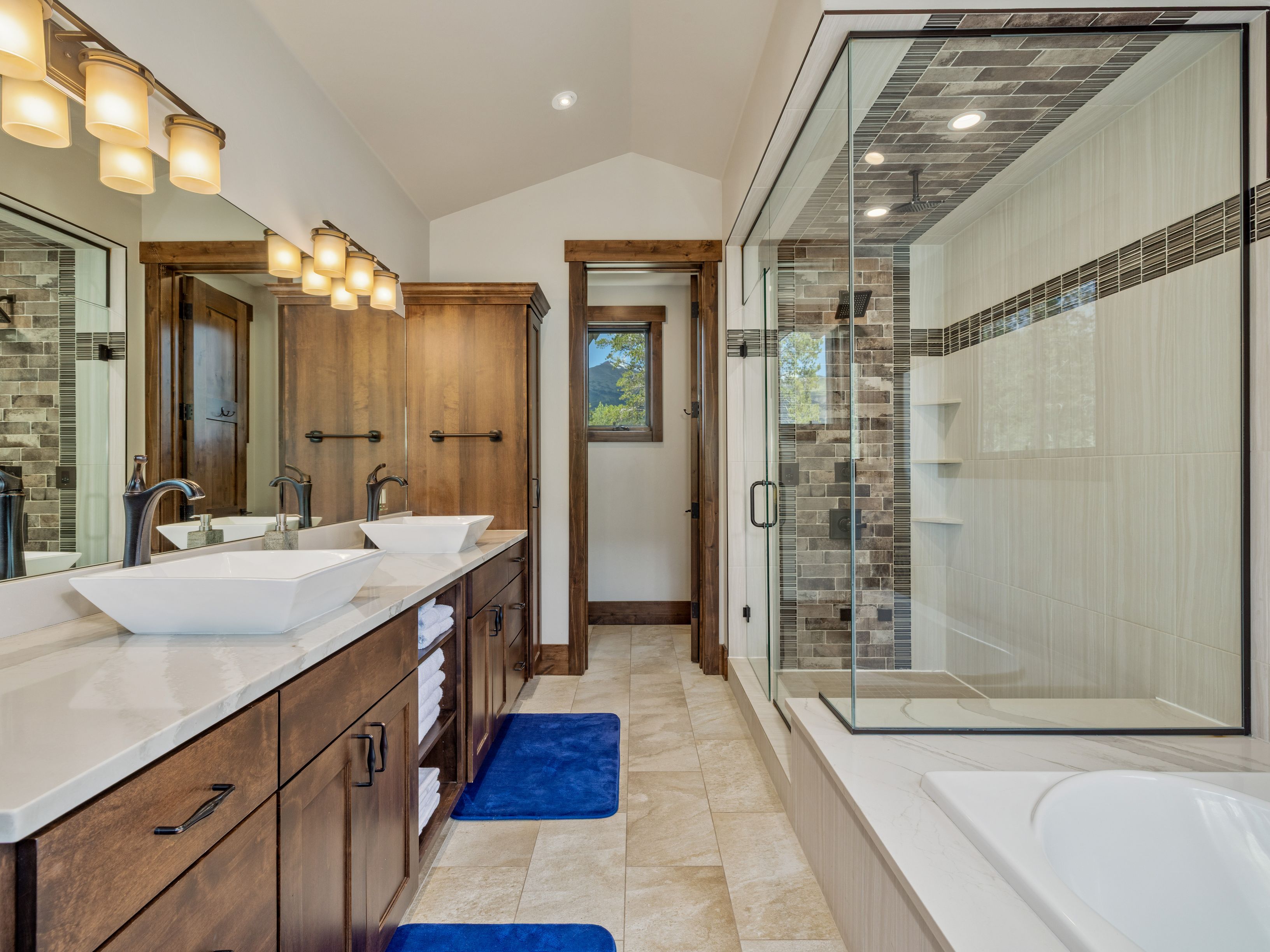 This primary bathroom features a dual-sink vanity with dark wood cabinetry, white vessel sinks, and a light-toned stone countertop. To the right, a glass-enclosed walk-in shower showcases a decorative tile accent wall, while a soaking tub is positioned in the foreground. The space is finished with neutral floor tiles and warm lighting, creating a clean and inviting atmosphere.