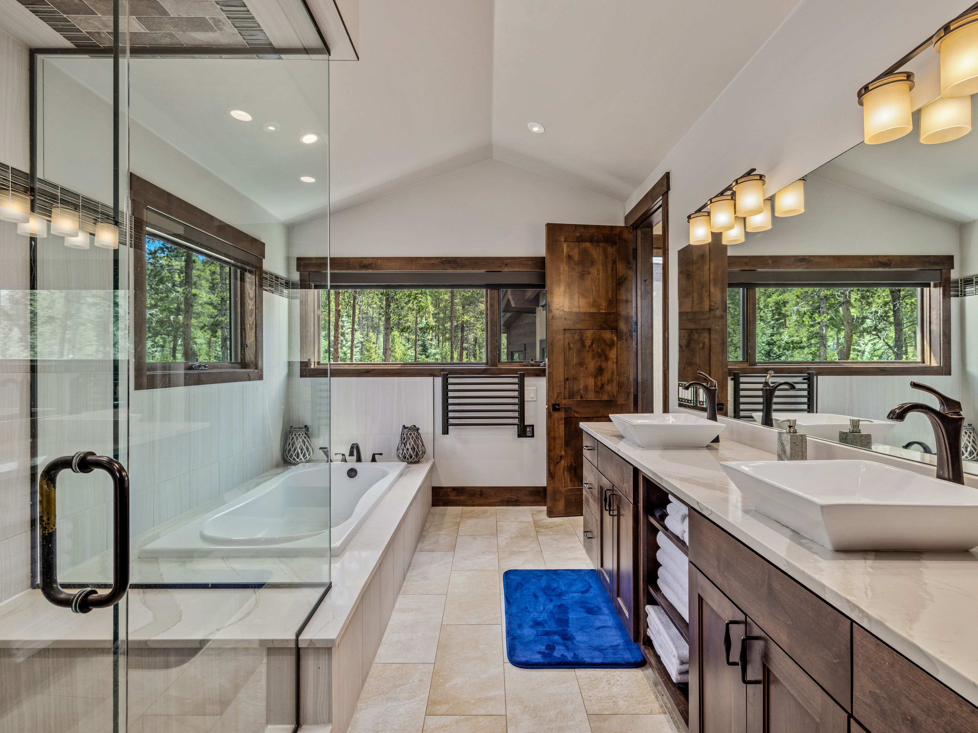 This primary bathroom features a rustic-modern aesthetic with dark wood cabinetry, dual vessel sinks, and a large soaking tub positioned beside a glass-enclosed shower. The space is illuminated by warm vanity lighting and natural light streaming through large windows that overlook a wooded area. A bright blue bath mat adds a pop of color to the neutral stone-tiled floor, creating a serene and inviting atmosphere.