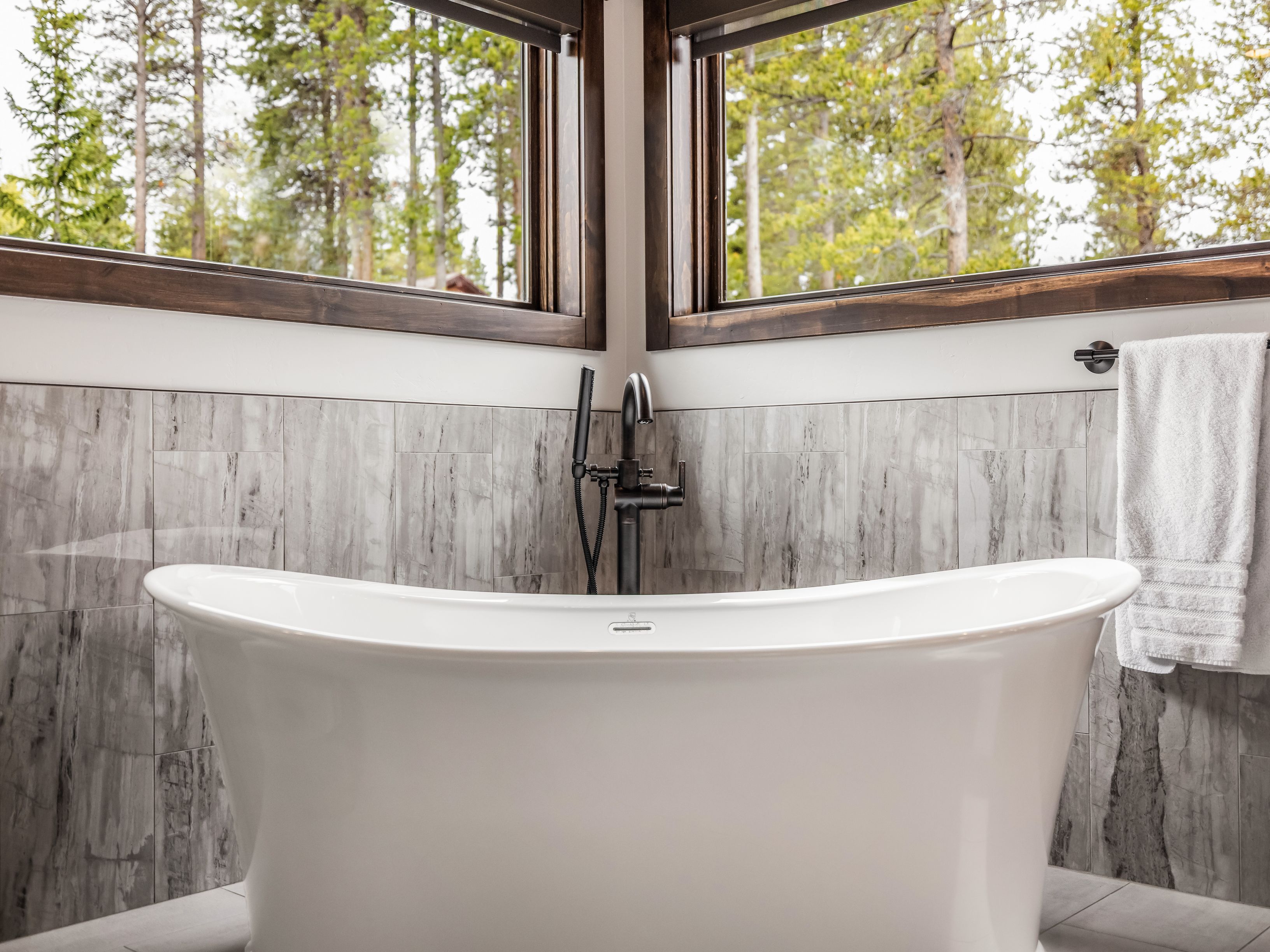 This primary bathroom features a sleek, white freestanding soaking tub positioned beneath a corner window arrangement that offers a serene view of the surrounding forest. The wall behind the tub is finished with vertical, grey-toned stone tiles, complemented by a dark metal faucet and handheld shower fixture. A white towel hangs on a wall-mounted rack to the right, contributing to the clean, modern, and tranquil aesthetic of the space.