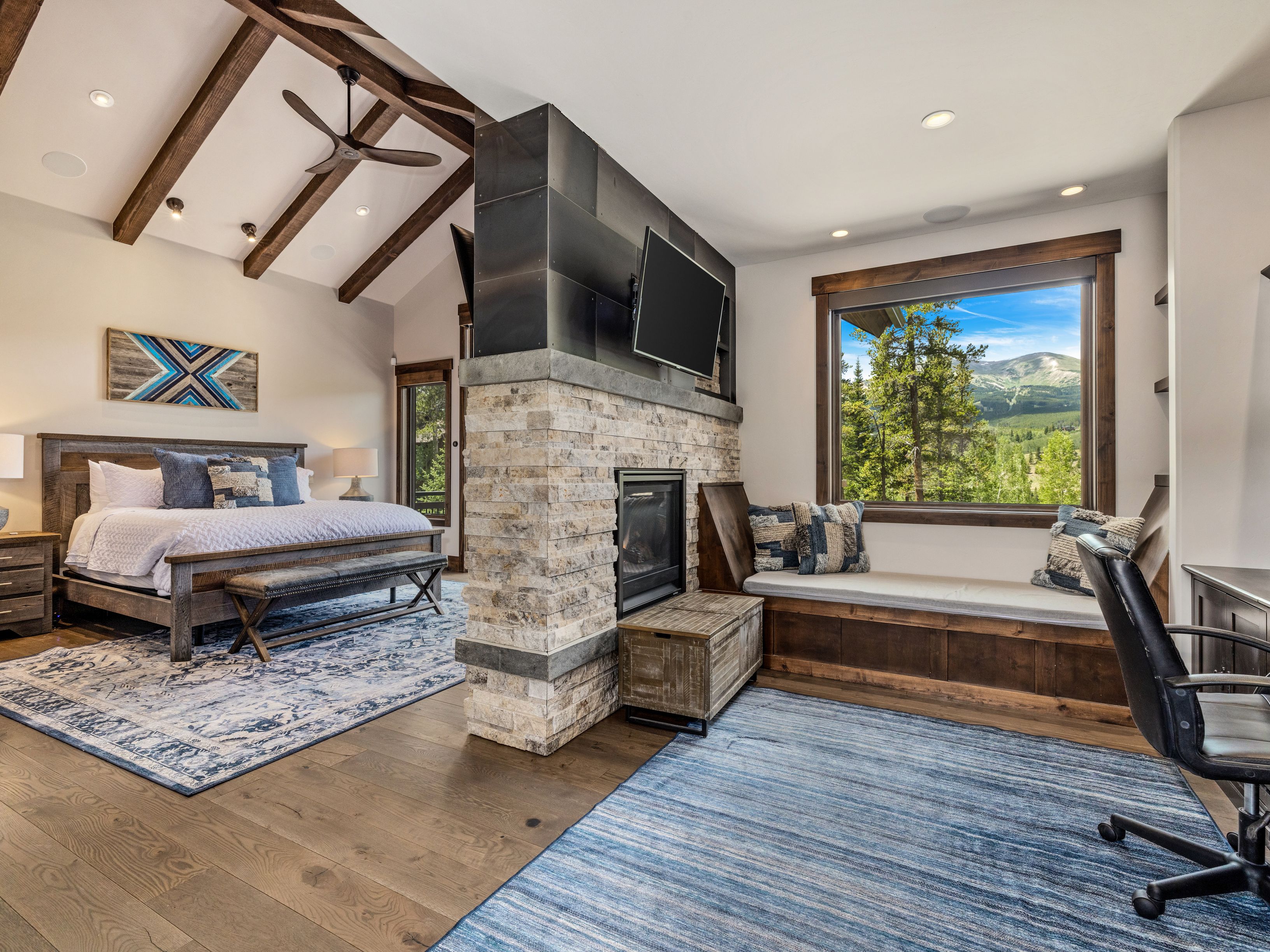 This spacious primary bedroom features a rustic, mountain-inspired aesthetic with exposed wooden ceiling beams and a central stone-clad chimney breast that separates the sleeping area from a cozy window nook. The room is anchored by a large bed with a patterned rug and includes a dedicated office space with a desk and chair, all bathed in natural light from a large window overlooking a scenic mountain landscape. The combination of warm wood tones, stone textures, and a neutral color palette creates a serene and inviting atmosphere.