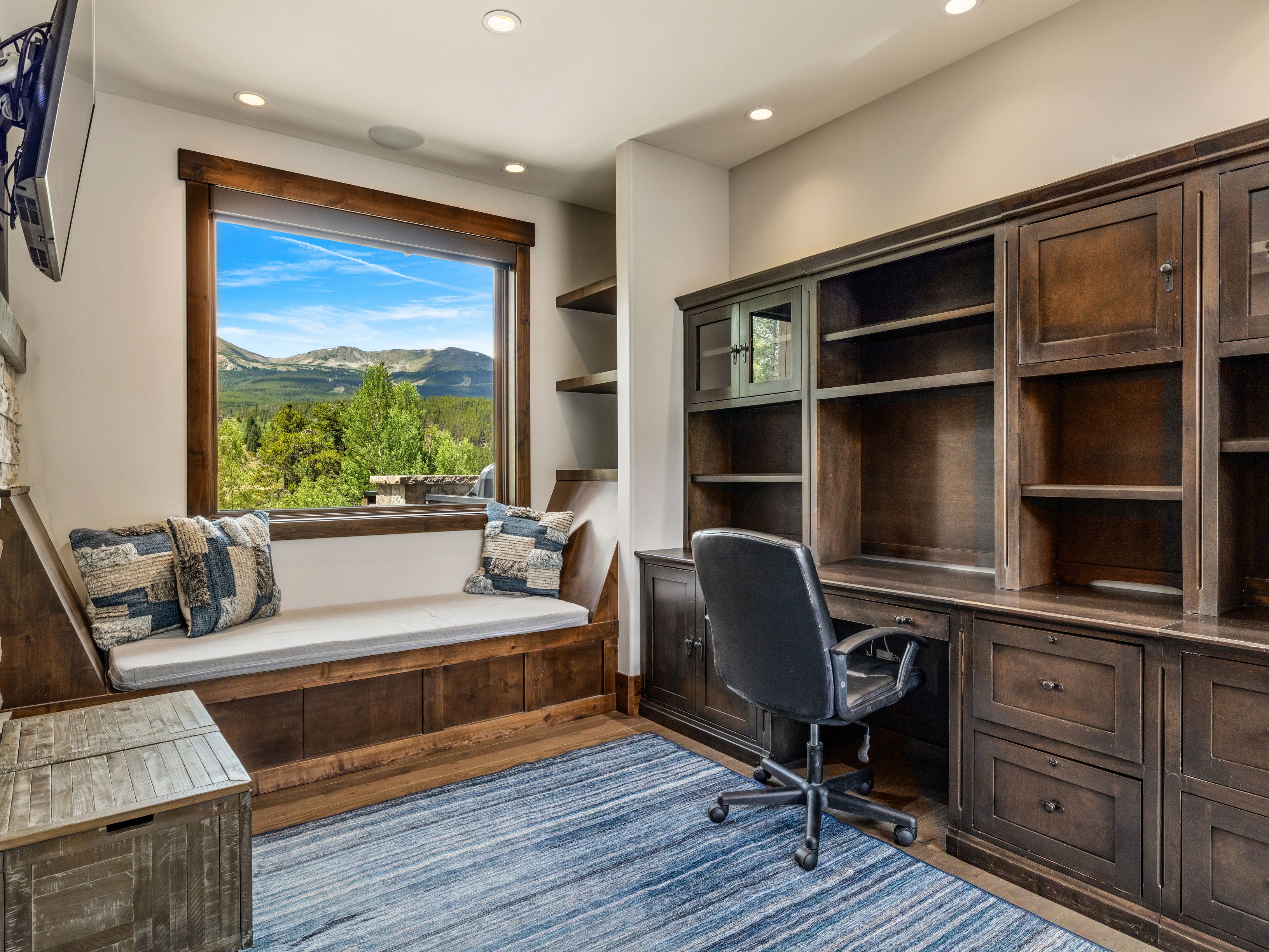 This home office features a built-in dark wood desk and shelving unit, providing ample storage and workspace. A cozy window seat with patterned cushions sits beneath a large window that frames a scenic mountain view. The room is completed by a blue-toned area rug and a black office chair, creating a professional yet comfortable atmosphere.