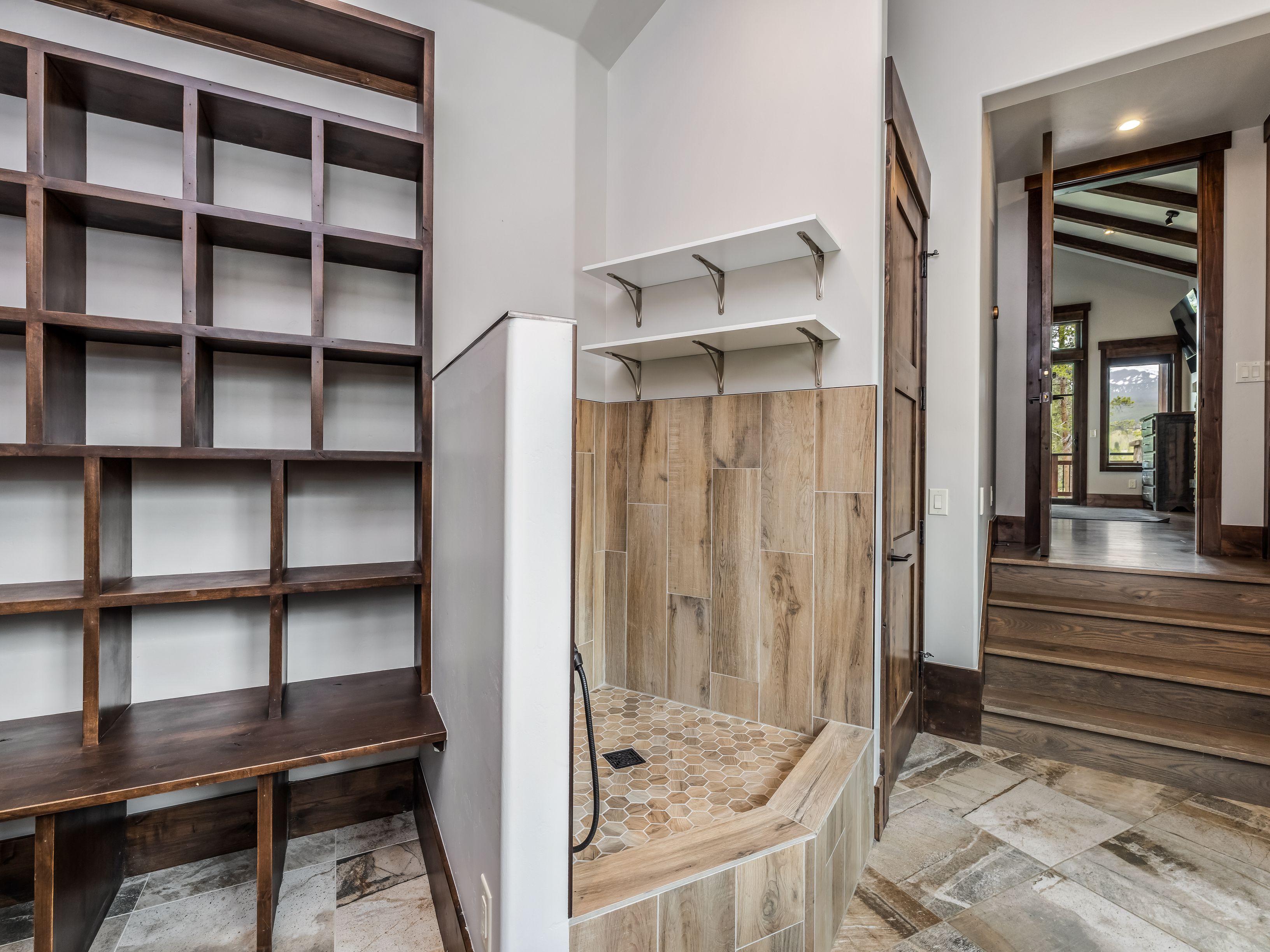 This interior space features a dedicated pet shower area with wood-look tile walls and a hexagonal tile floor, positioned next to a custom dark wood shelving unit. A set of white floating shelves is mounted above the shower, and a wooden door leads to an adjacent room with stairs. The overall aesthetic is rustic and functional, utilizing natural textures and warm wood tones throughout the space.