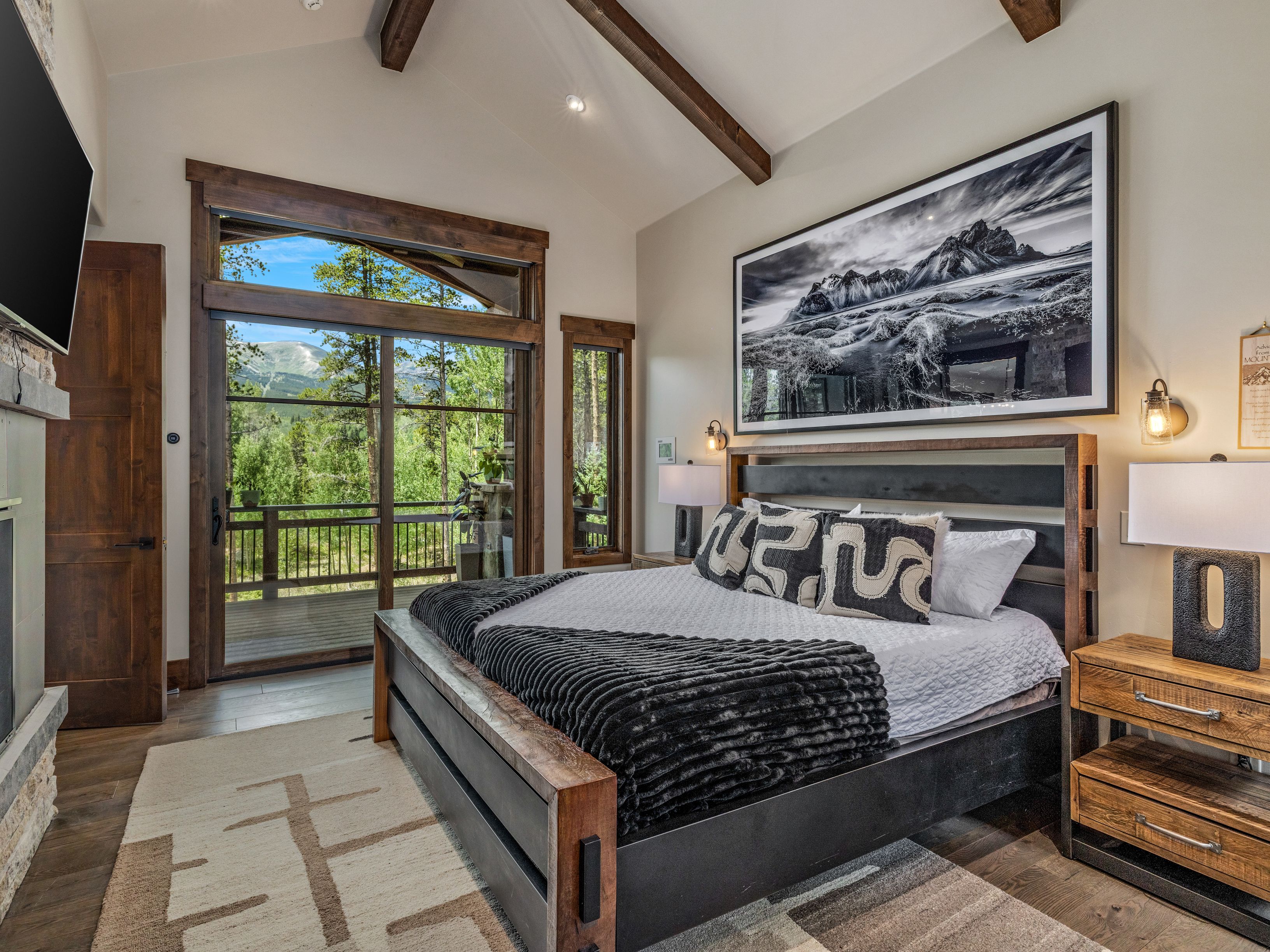 This primary bedroom features a rustic-modern aesthetic with a large bed frame, a textured black throw, and patterned accent pillows. A prominent black-and-white landscape photograph hangs above the headboard, while a sliding glass door provides access to a deck with mountain views. The room is accented by warm wood trim, exposed ceiling beams, and bedside lamps, creating a cozy and inviting atmosphere.
