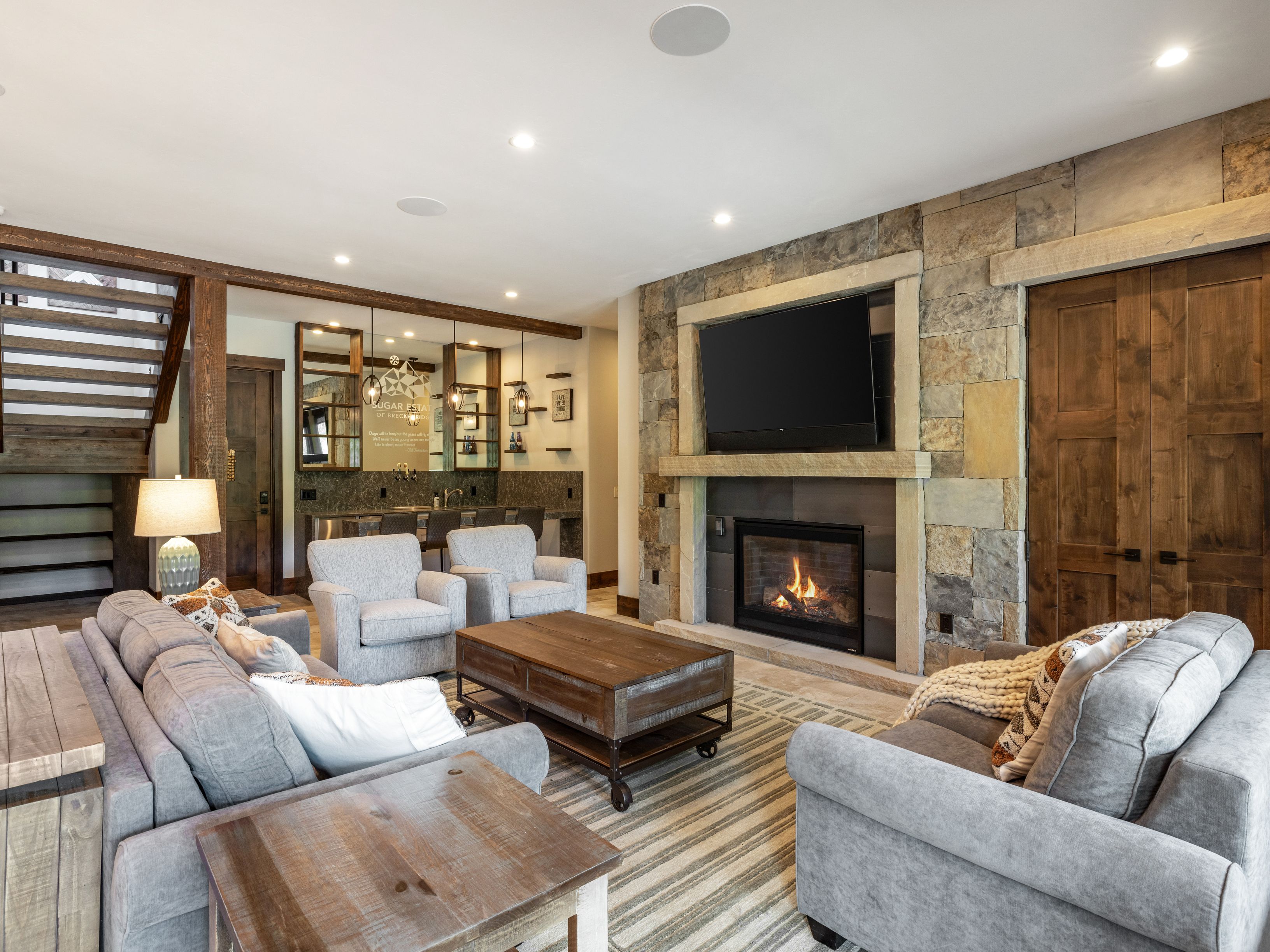 This inviting living room features a prominent stone-clad chimney breast housing a fireplace and a mounted television. The space is furnished with plush gray seating, a rustic wooden coffee table, and a striped area rug, all set against a backdrop of warm wood tones and a staircase to the left. A wet bar area with glass shelving and pendant lighting is visible in the background, creating a cohesive and cozy atmosphere perfect for entertaining.