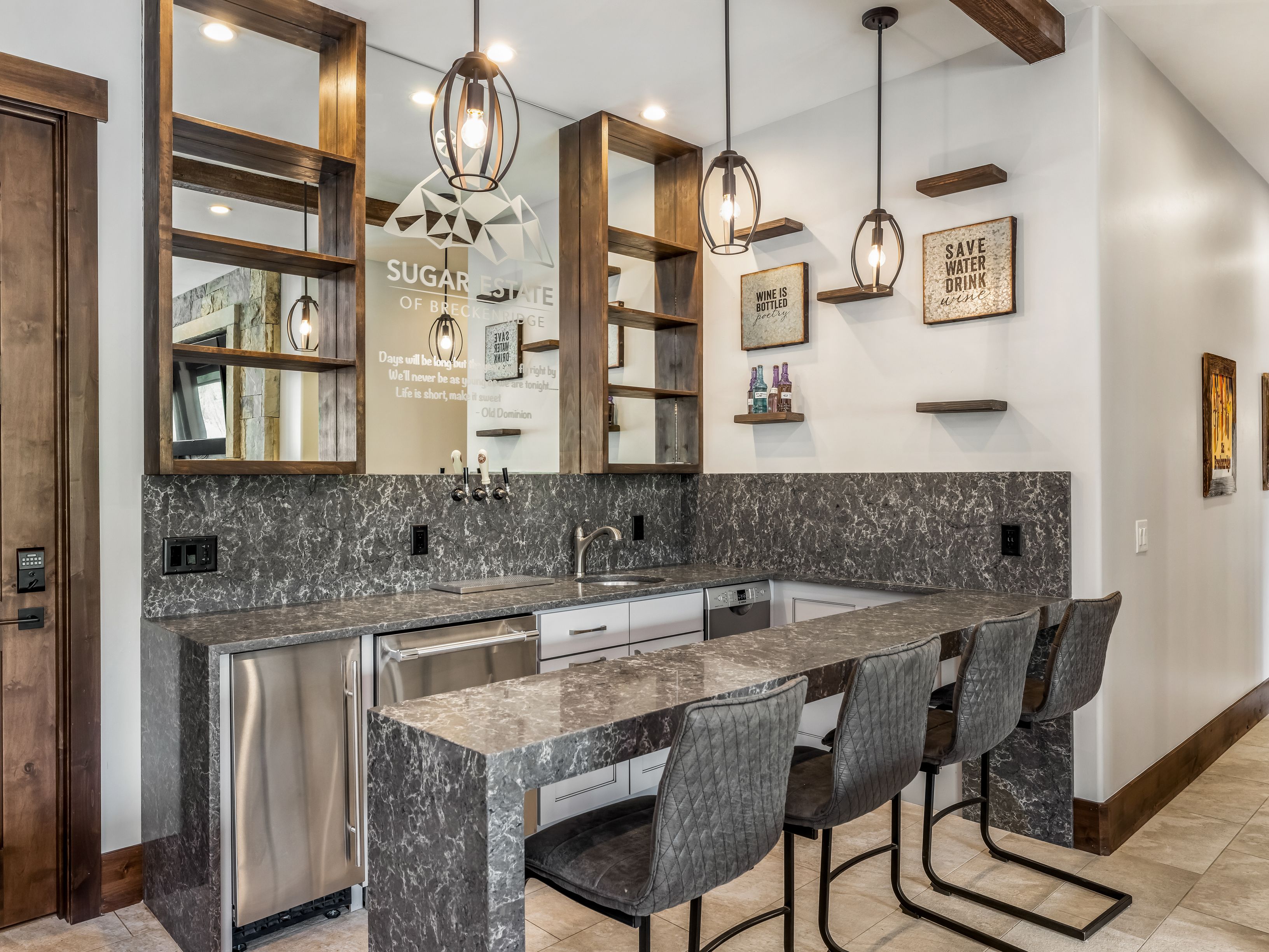 This stylish home bar area features a dark, patterned stone countertop and matching backsplash, complemented by white cabinetry and open wood shelving. The space is illuminated by three industrial-style pendant lights and includes a built-in stainless steel beverage refrigerator and sink. Four modern, quilted-leather bar stools are tucked under the extended counter, creating an inviting atmosphere for entertaining.