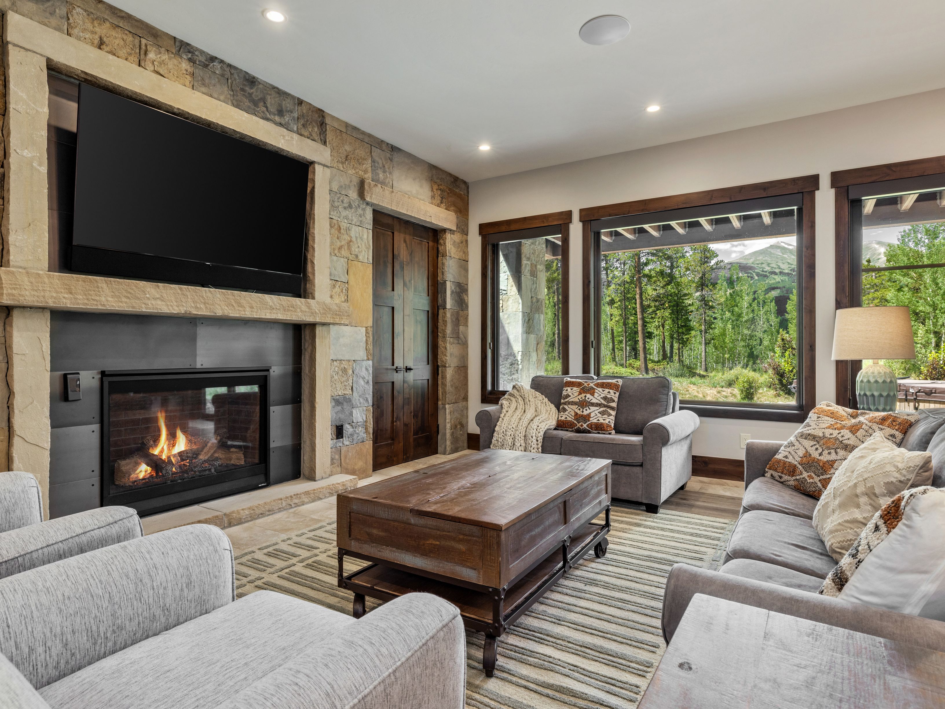 This inviting living room features a prominent stone-clad chimney breast surrounding a fireplace, complemented by a large television mounted above. The space is furnished with plush gray seating and a rustic wooden coffee table, all arranged on a textured area rug. Large windows provide a scenic view of the surrounding forest and mountains, creating a bright and tranquil atmosphere.