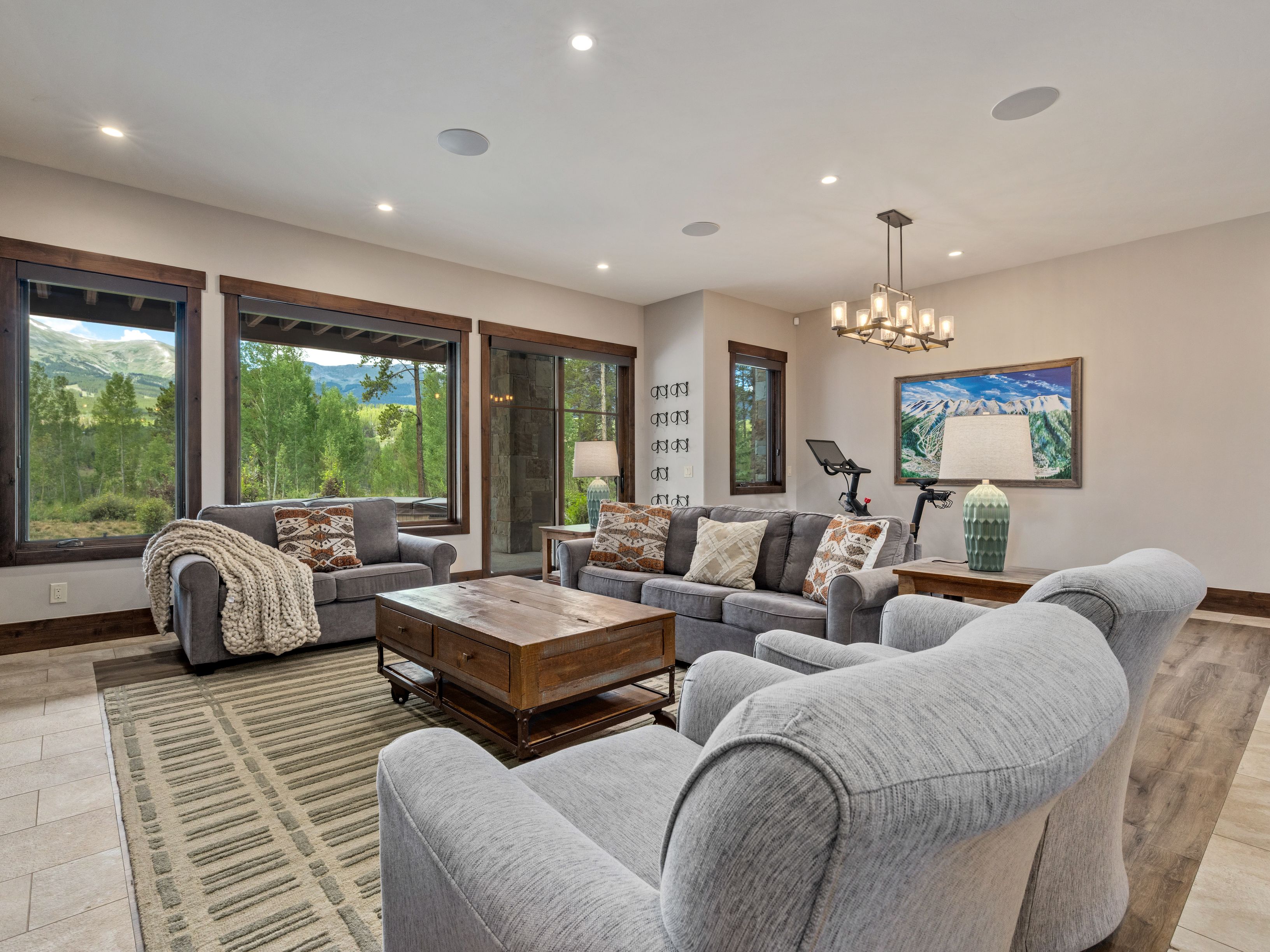 This spacious living room features a comfortable seating arrangement with a large gray sofa, a matching loveseat, and two armchairs centered around a rustic wooden coffee table. Large windows provide expansive views of the surrounding forest and mountains, filling the room with natural light. The space is accented by a patterned area rug, a decorative chandelier, and a framed landscape print, creating a warm and inviting atmosphere.