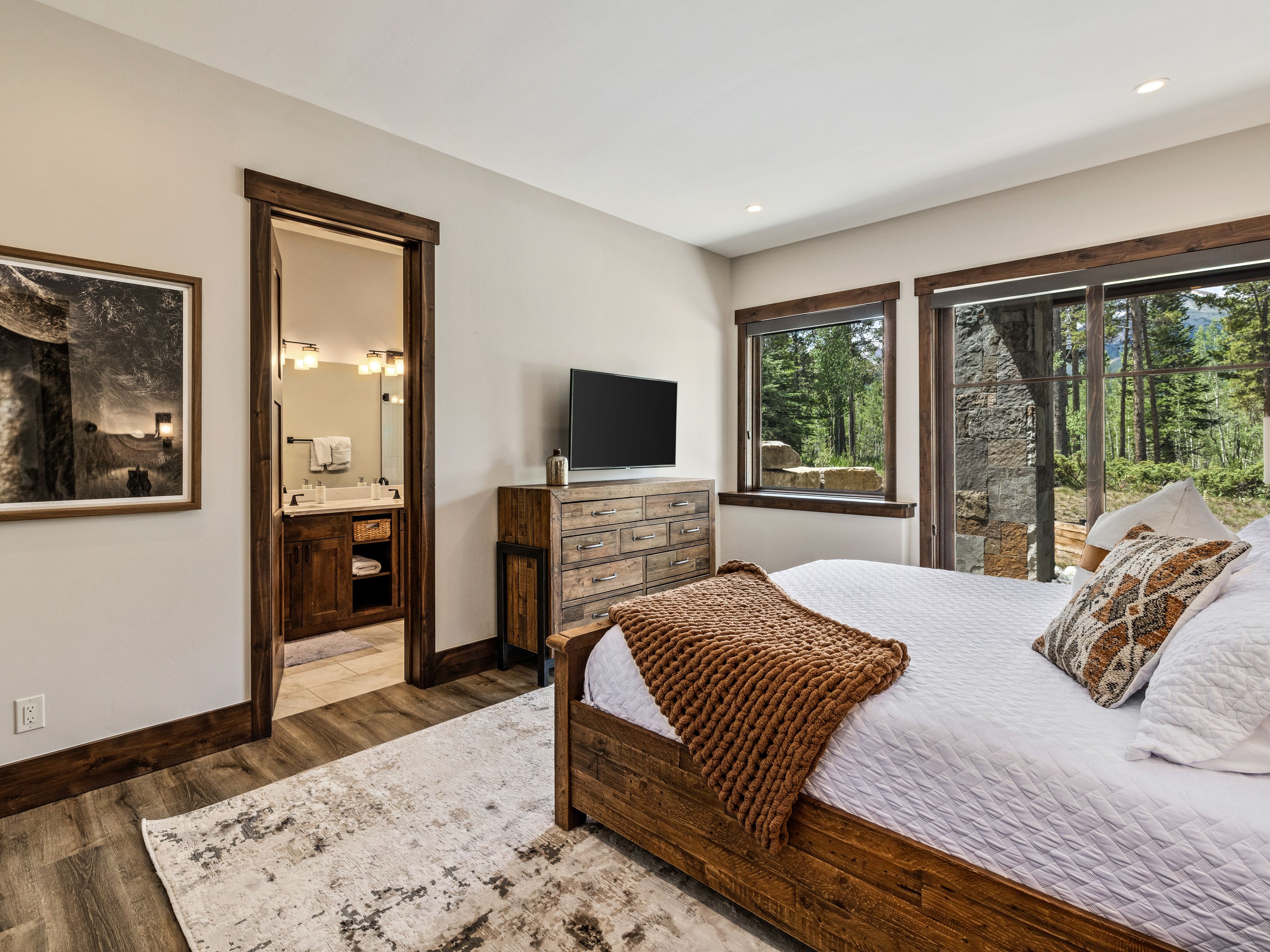 This primary bedroom features a rustic aesthetic with warm wood trim, a wooden bed frame, and a textured brown throw blanket. A dresser sits against the wall beneath a wall-mounted television, while a doorway leads into an adjoining bathroom. Large windows provide a view of the surrounding forest, and a patterned area rug anchors the space on the wood-look flooring.