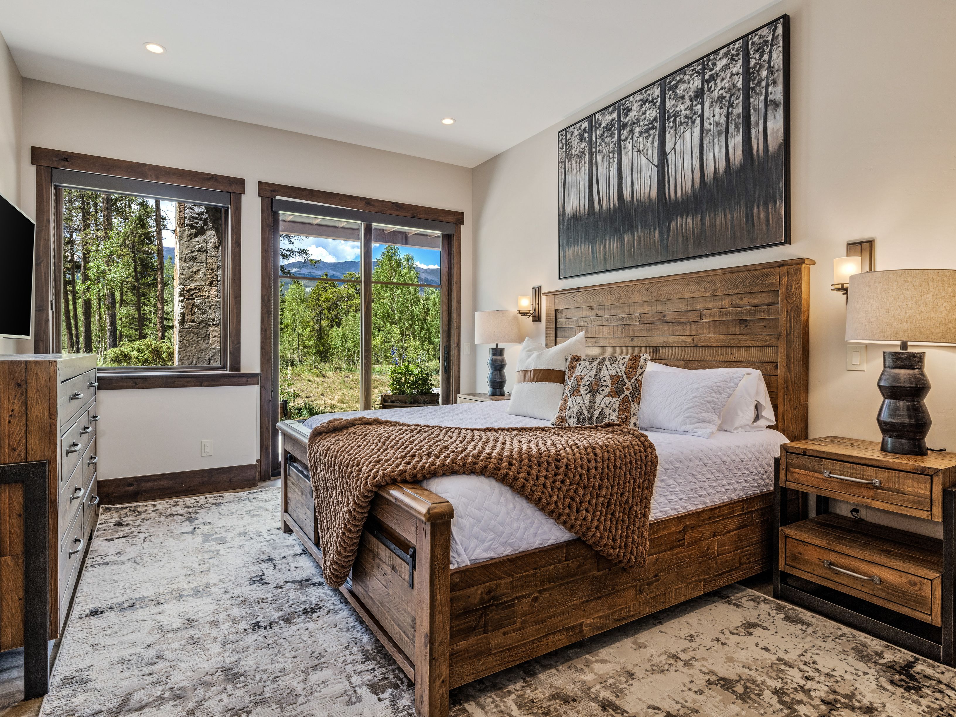This primary bedroom features a rustic aesthetic with a substantial wood-framed bed, a chunky knit throw blanket, and matching wood nightstands. A large piece of forest-themed wall art hangs above the headboard, while a sliding glass door provides a view of the surrounding trees and mountains. The room is grounded by a neutral, patterned area rug and illuminated by warm bedside lamps.