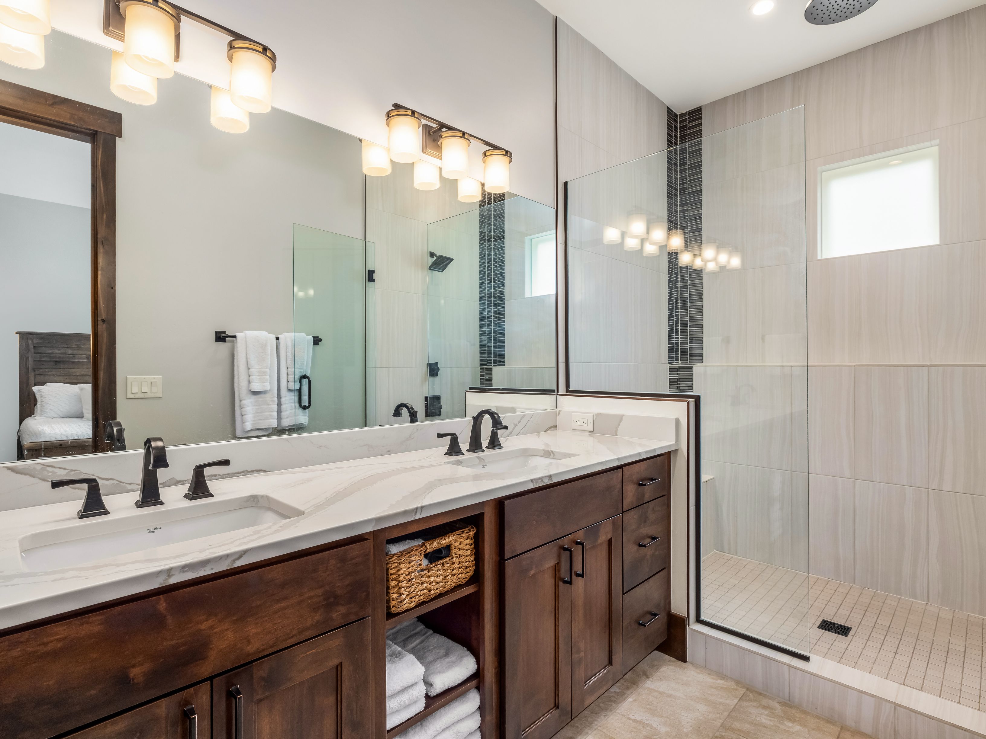 This primary bathroom features a dual-sink vanity with dark wood cabinetry and a light-toned stone countertop. A large mirror spans the wall above the vanity, reflecting the adjacent walk-in shower which is enclosed by glass panels and finished with light-colored wall tiles. The space is well-lit by two multi-bulb vanity lights, creating a clean and functional aesthetic.