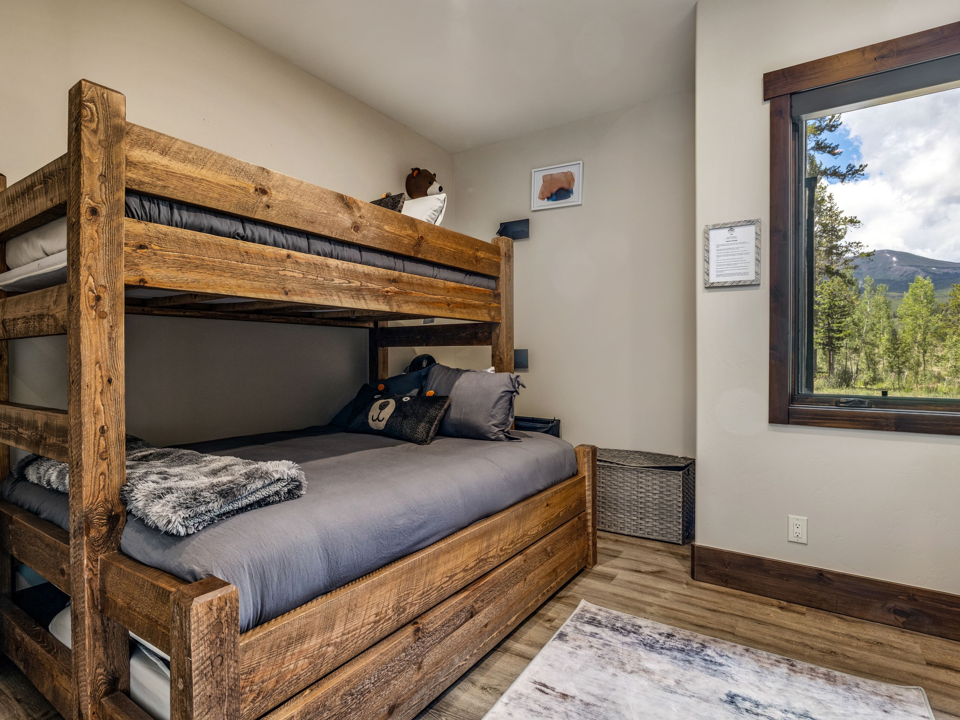This cozy bedroom features a rustic, heavy-timber bunk bed set with dark gray bedding, creating a warm and inviting atmosphere. A large window to the right provides a scenic view of the surrounding forest and mountains, while a woven storage bin sits in the corner. The room is finished with light-toned wood flooring and neutral walls, emphasizing a comfortable, cabin-inspired aesthetic.
