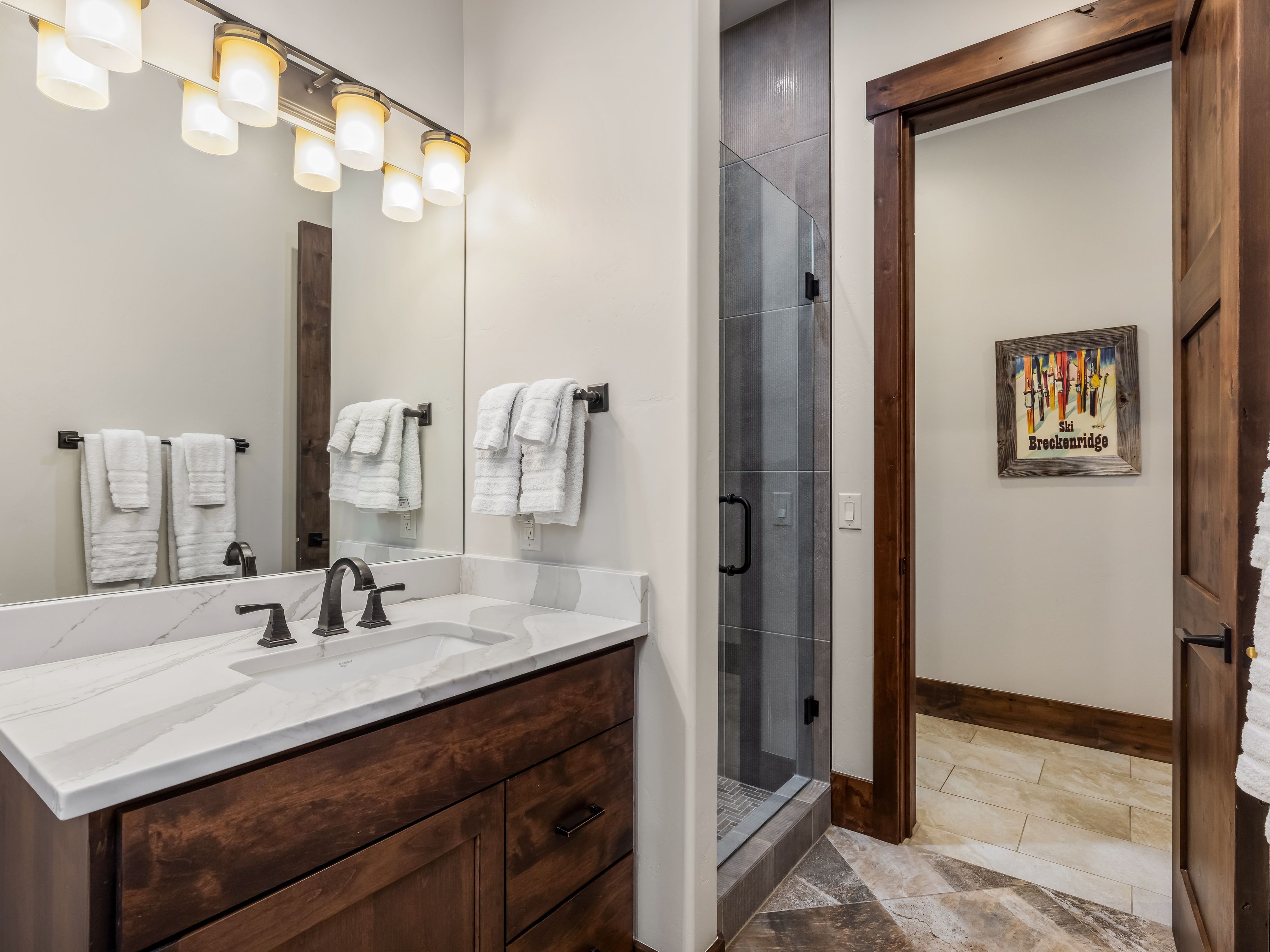 This bathroom features a dark wood vanity topped with a light-colored stone counter and a modern faucet. A large mirror reflects the vanity and a wall-mounted light fixture, while a glass-enclosed shower with dark tile is positioned to the right. A doorway leads into an adjacent room decorated with a framed ski-themed print, creating a cohesive, rustic-inspired aesthetic.