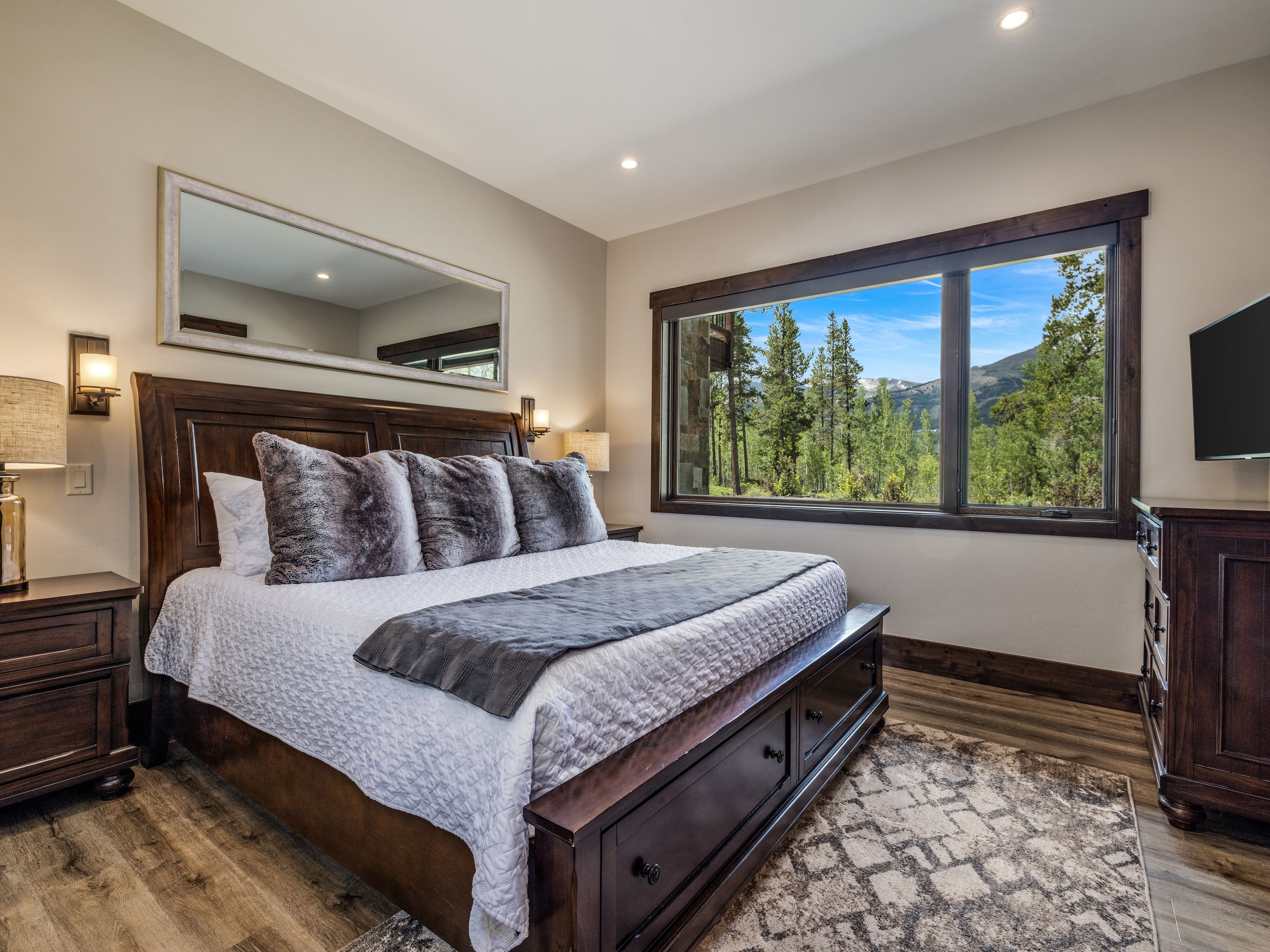 This primary bedroom features a rich, dark wood bed frame with integrated storage drawers and a plush white quilt topped with textured gray pillows. A large window provides a scenic view of the surrounding forest and mountains, while a wide mirror above the headboard enhances the sense of space. The room is finished with wood-look flooring, a patterned area rug, and matching dark wood bedside tables, creating a cozy and rustic mountain retreat atmosphere.