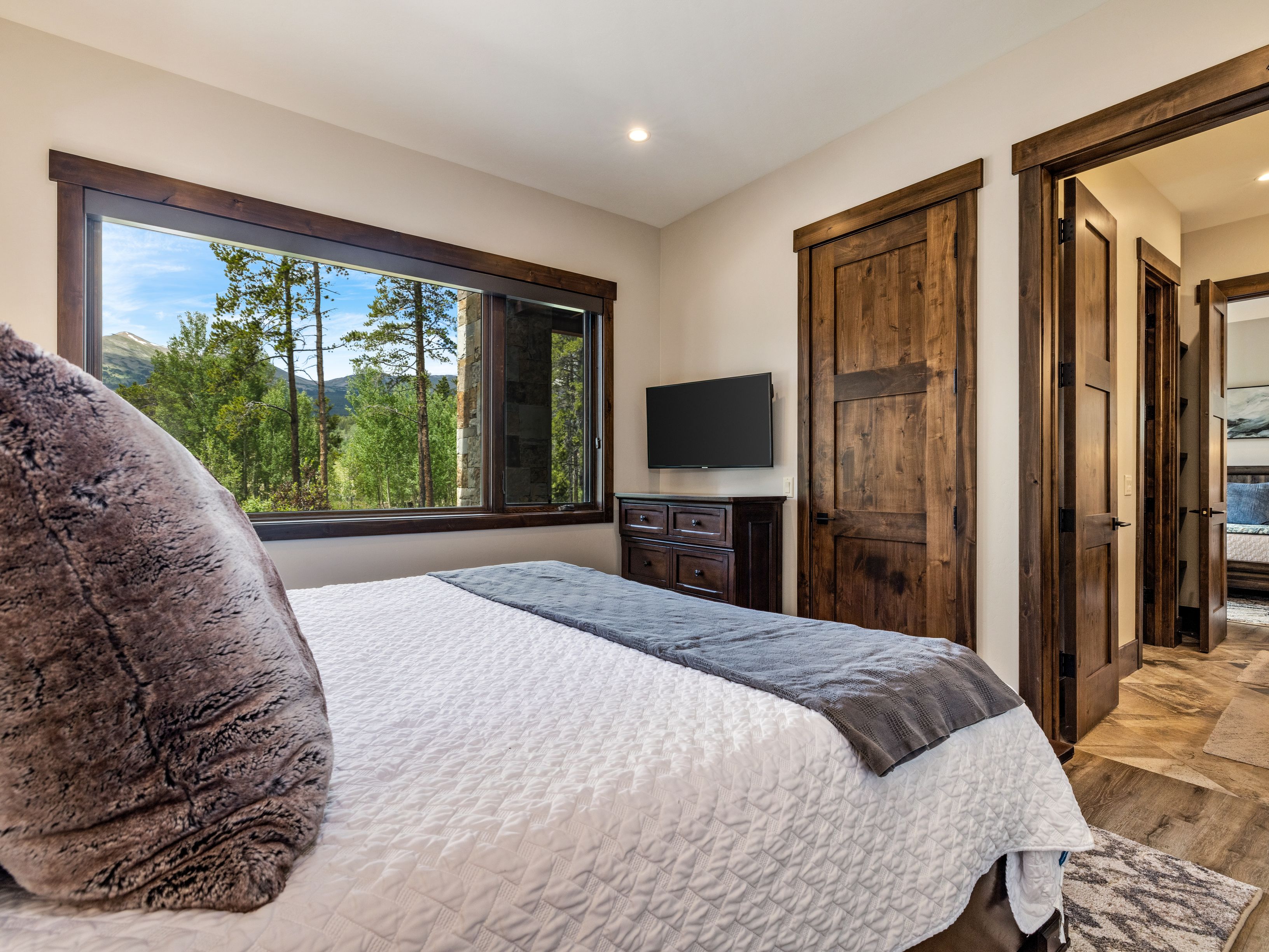 This cozy bedroom features a plush bed with a white quilted coverlet and a dark gray runner, accented by a large, textured throw pillow. A wide window with dark wood trim provides a scenic view of the surrounding forest and mountains, while a dark wood dresser sits below a wall-mounted television. The room transitions into a hallway with matching dark wood doors, showcasing a cohesive rustic aesthetic with warm, neutral wall tones.